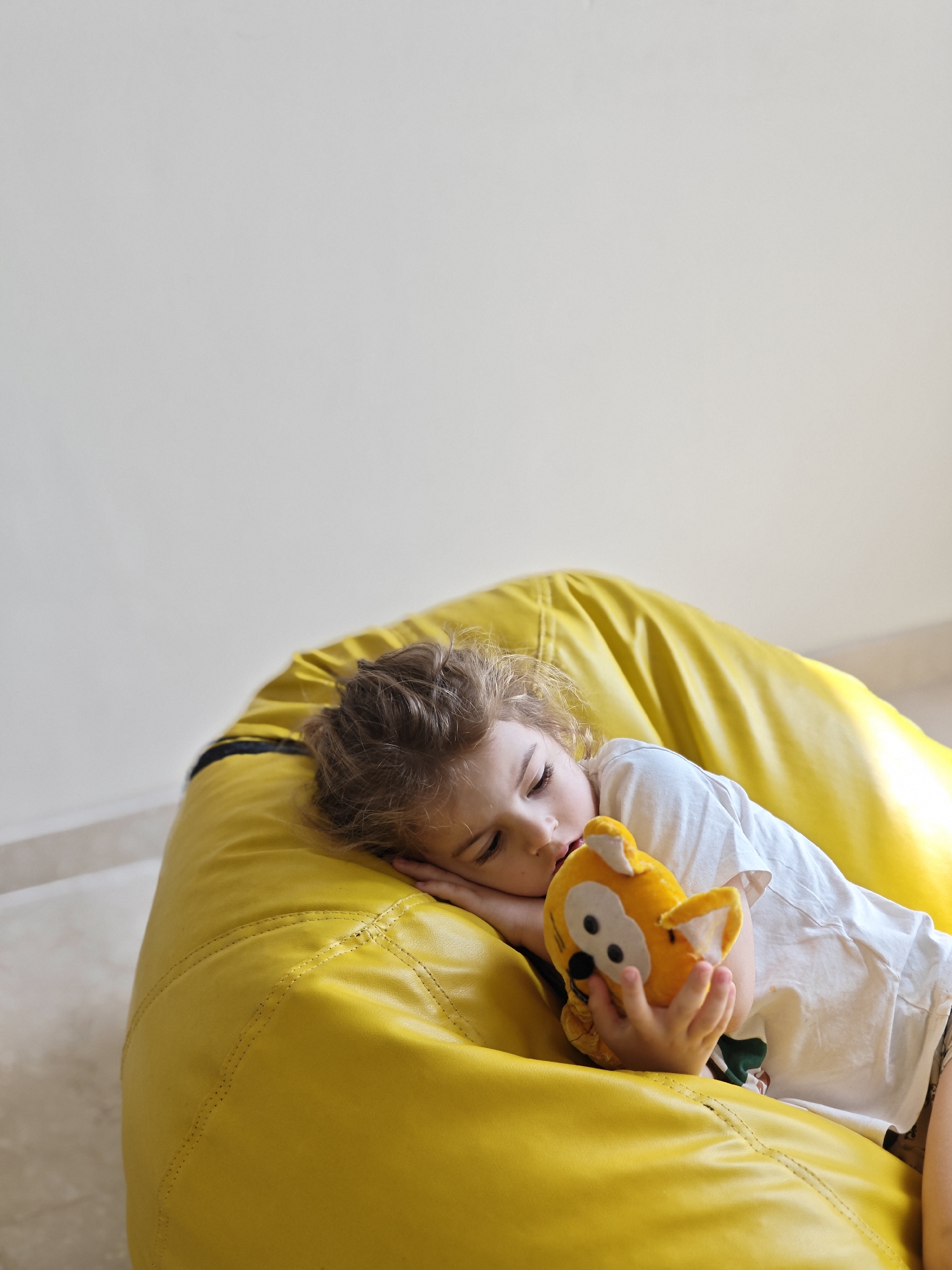 A young child is lying on a large, yellow beanbag chair indoors. The child is light-skinned with curly hair, wearing a white t-shirt. They are holding a yellow plush toy, possibly a fox or similar animal, close to their face with one hand. The beanbag chair appears to be made of leather or a similar material. The background consists of a plain, off-white wall and a floor with light-colored tiling. No information indicates the location is Mumbai, India.