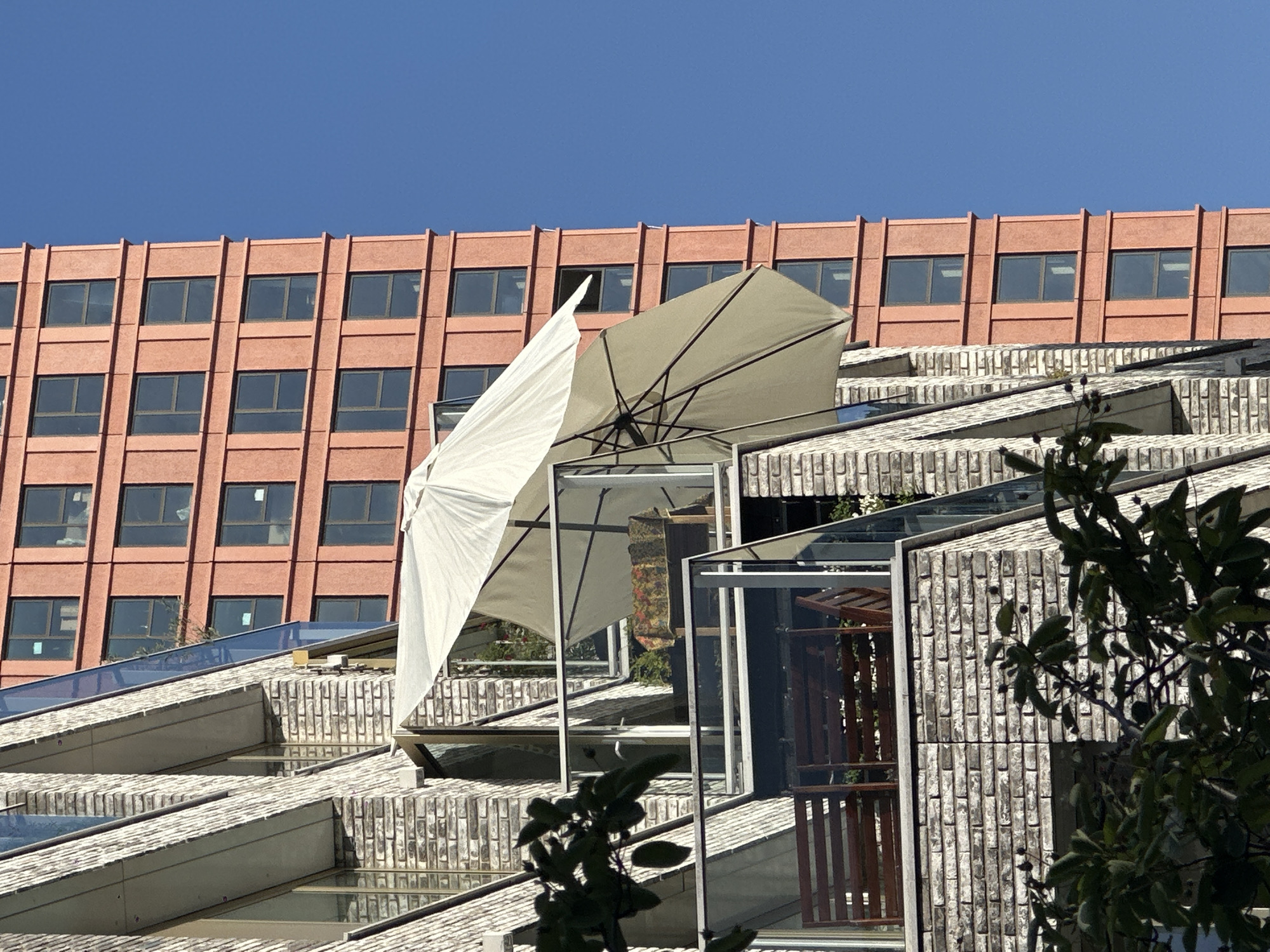The image shows a building with an unusual stepped or terraced roof structure, and an orange building behind it. The stepped roofs are constructed of brick and glass panels. A large, white, open umbrella is perched precariously on one of the steps. The building behind has a grid of windows. Green foliage is visible in the foreground, partially obscuring the scene. This is likely the "Kaupenzaal" building in Amsterdam, Netherlands.