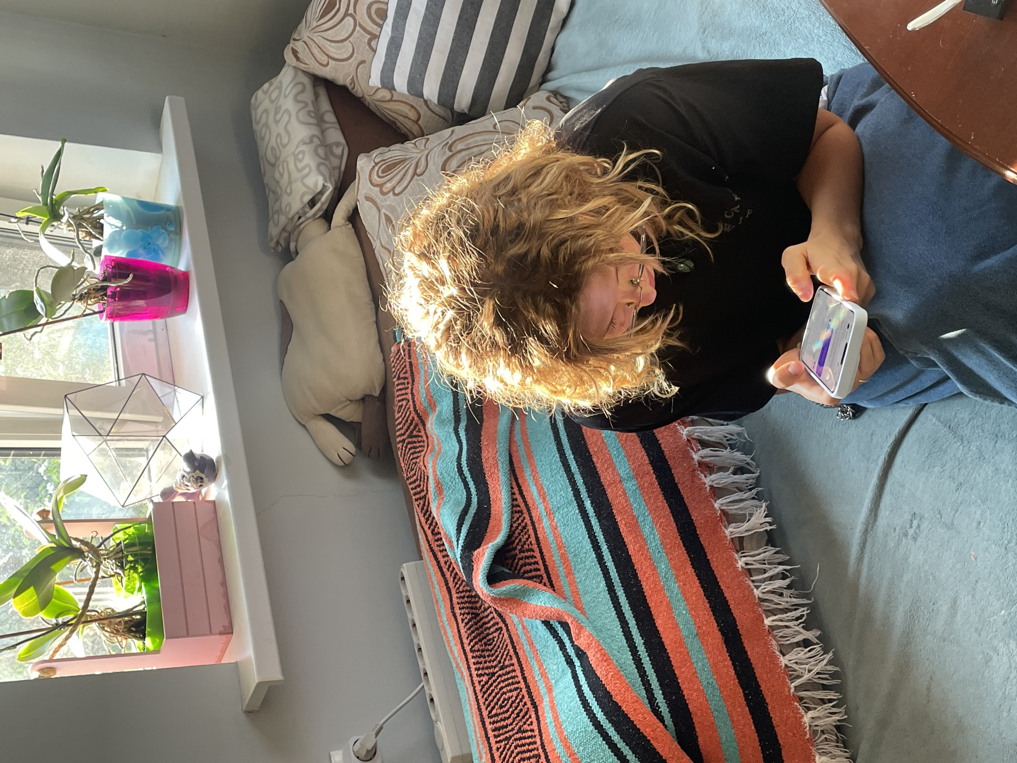 A person with curly blonde hair, wearing glasses and a black shirt, is lying on a blue bed or couch, looking at a smartphone held in their hands. The background includes a striped blanket with orange, black, and blue stripes, patterned pillows, and a windowsill with plants in colorful pots. The location appears to be indoors. There is not enough information to determine if this image was taken in Sestroretsk, Russia.