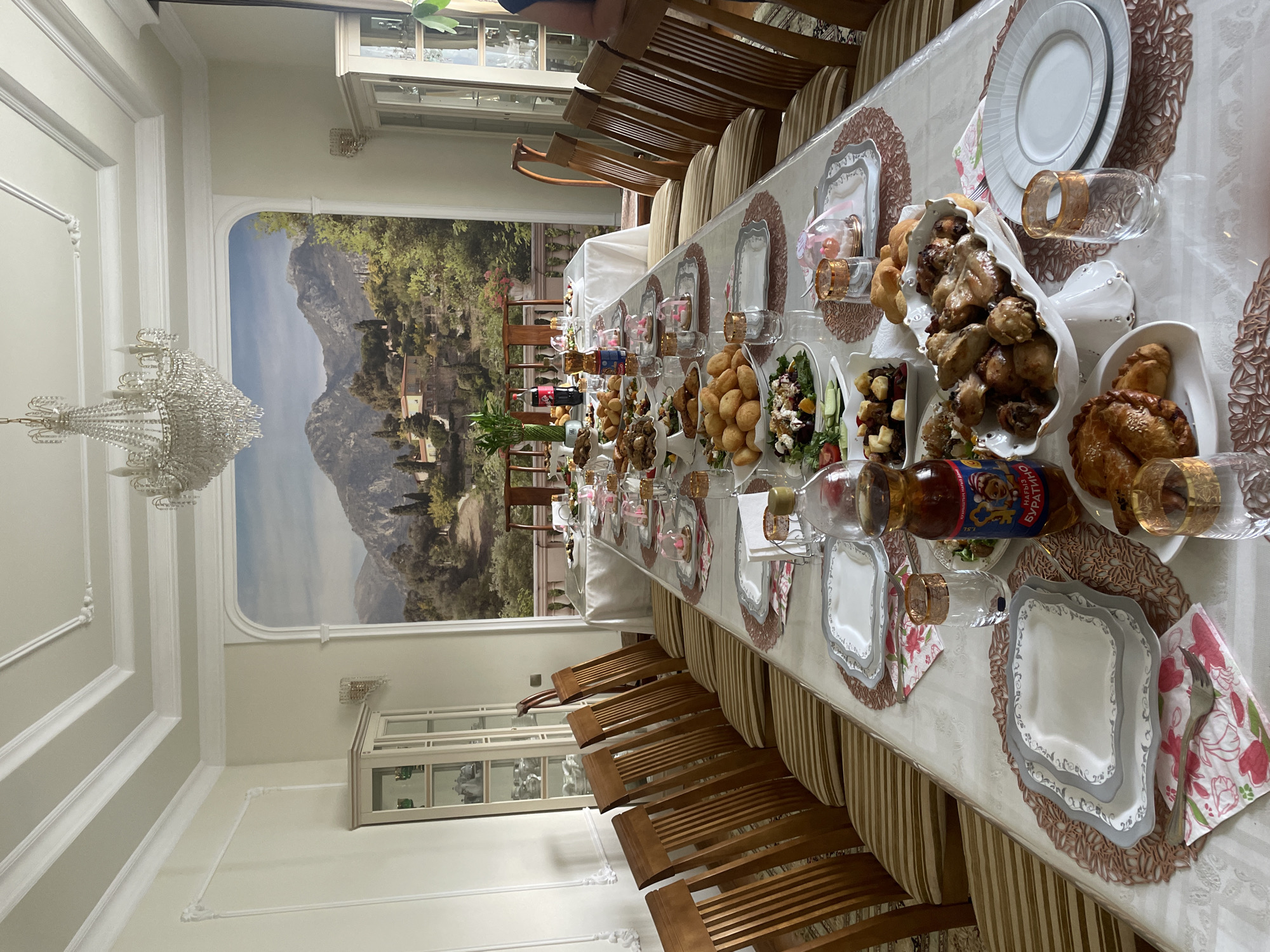 The image shows a formal dining room set for a meal. A long table is laden with a variety of dishes, including roasted chicken, pastries, and salads. Place settings with plates, glasses, and silverware are neatly arranged for multiple guests. The room is decorated with ornate moldings on the ceiling and walls, and a large mural depicting a mountainous landscape adorns one wall. A crystal chandelier hangs from the ceiling. The setting suggests a celebratory occasion or a formal gathering. Based on the prominent display of the "Буратино" (Buratino) beverage bottle, which is a popular Russian and Kazakh soda, and the general decor, the scene likely takes place in Almaty, Kazakhstan. The lighting suggests it is daytime. No individuals are clearly visible in a way that identifies specific actions or interactions.