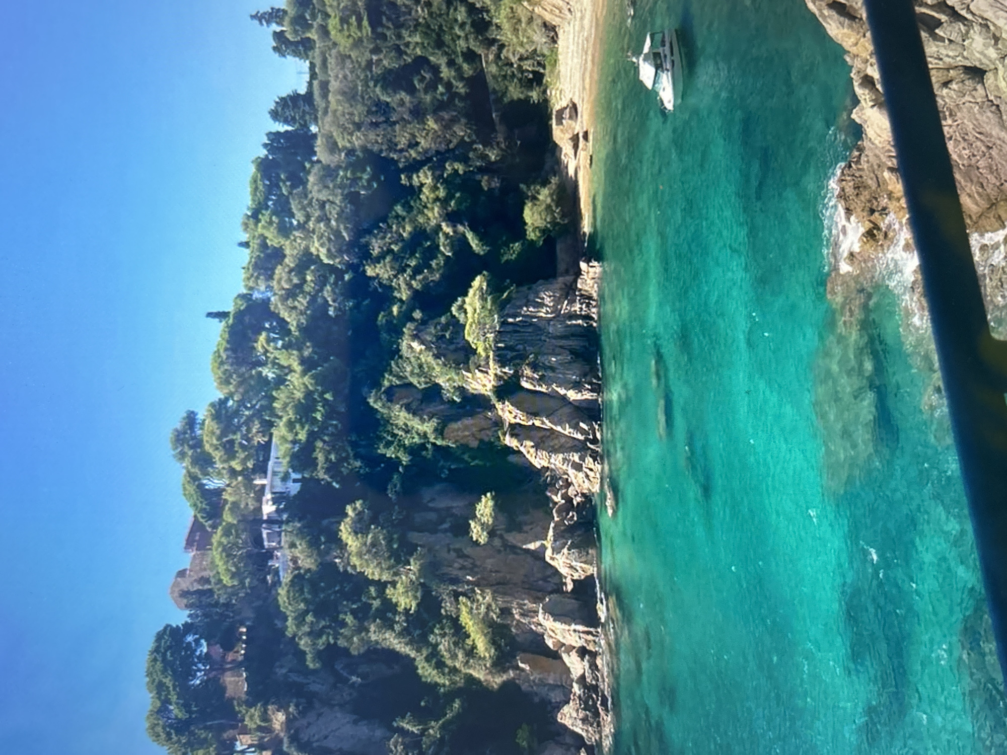 The image displays a picturesque coastal landscape under a bright, clear blue sky. A lush green hillside, densely covered with trees and Mediterranean-type vegetation, dominates the left side, sloping down towards the sea. Scattered among the trees on the hillside are several white buildings, suggesting a secluded settlement or villas.
The coastline is rugged, featuring prominent rocky cliffs and outcrops that meet the remarkably clear, turquoise-blue water. The transparency of the water allows for views of the seabed in certain areas. Along the shore, small, secluded coves with patches of sand are visible. In one such cove on the right side of the image, a white motorboat with a cabin is anchored in the calm water, positioned near a small sandy beach.
A dark, vertical object, possibly a pole or part of a railing from where the photo was taken, partially obstructs the view on the far right edge. The scene conveys a sense of tranquility and natural beauty, characteristic of a sunny day in a coastal region. No people are visible, and there is no discernible text in the image. The time of day appears to be daytime, with ample sunlight.