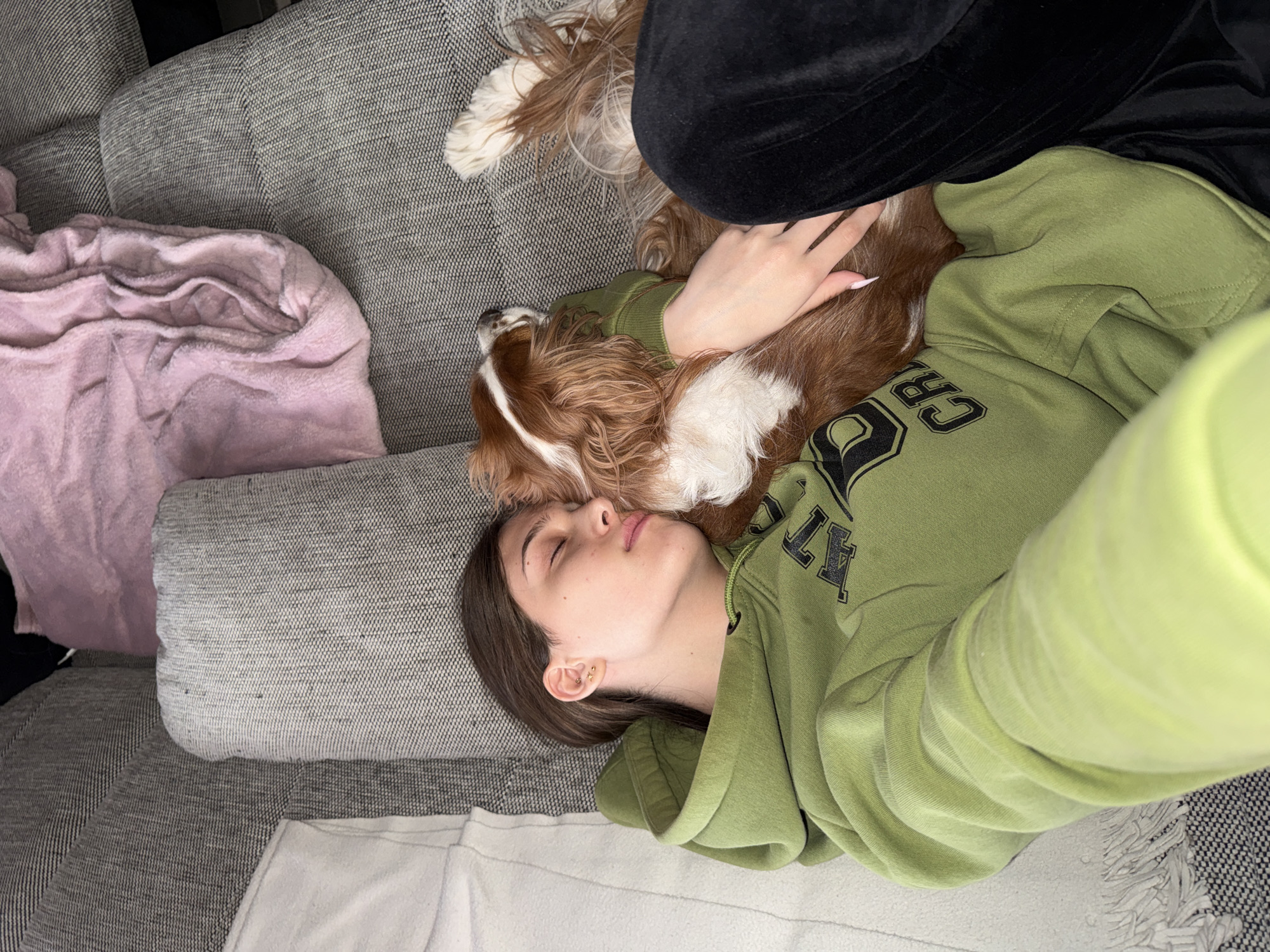 A person is lying on a gray couch with a Cavalier King Charles Spaniel dog resting on their chest. The person is wearing a green sweatshirt with black lettering. Their eyes are closed, and they appear to be sleeping. The dog is brown and white. A pink blanket is partially visible on the left side of the couch. A black object is near the dog, and a white blanket is visible at the bottom of the image. The location is unknown and there is no evidence to suggest it is in Sofia, Bulgaria.