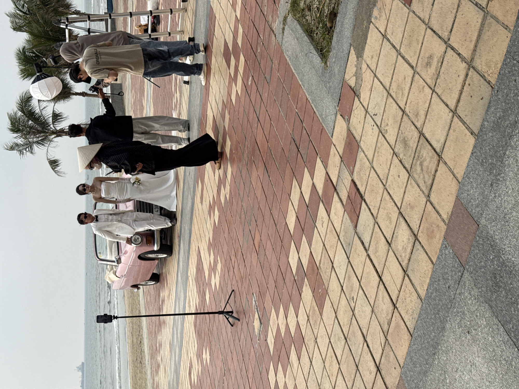 An outdoor wedding photoshoot is underway on a paved promenade adjacent to a beach in Da Nang, Vietnam. The ground is paved with a checkerboard pattern of alternating light tan and reddish-brown square tiles, bordered by a strip of grey granite.
In the mid-ground, a bridal couple poses beside a vintage light pink convertible car with its soft top down; a Vietnamese license plate beginning with "43A" is partially visible. The groom wears a white suit, white shirt, black bow tie, and sunglasses, leaning against the car's front fender. The bride, in a strapless white gown and sunglasses, holds a small white floral bouquet.
To the right of the couple, a four-person crew manages the photoshoot. One individual in a beige t-shirt and cap operates a professional camera. Another person in a black shirt holds equipment, facing the couple. A third individual, wearing a grey hooded sweatshirt, stands next to a silver folding ladder and a white softbox light diffuser mounted on a stand. A woman, possibly directing, wears a traditional Vietnamese conical hat (Nón Lá), sunglasses, a face mask pulled down, and a black outfit, holding a smartphone.
To the left of the couple, a smartphone is mounted on a tall black tripod, facing them. The background features a sandy beach leading to the ocean with visible waves. Several palm trees stand further back, and a large cargo ship is anchored or moving on the distant horizon under an overcast sky.