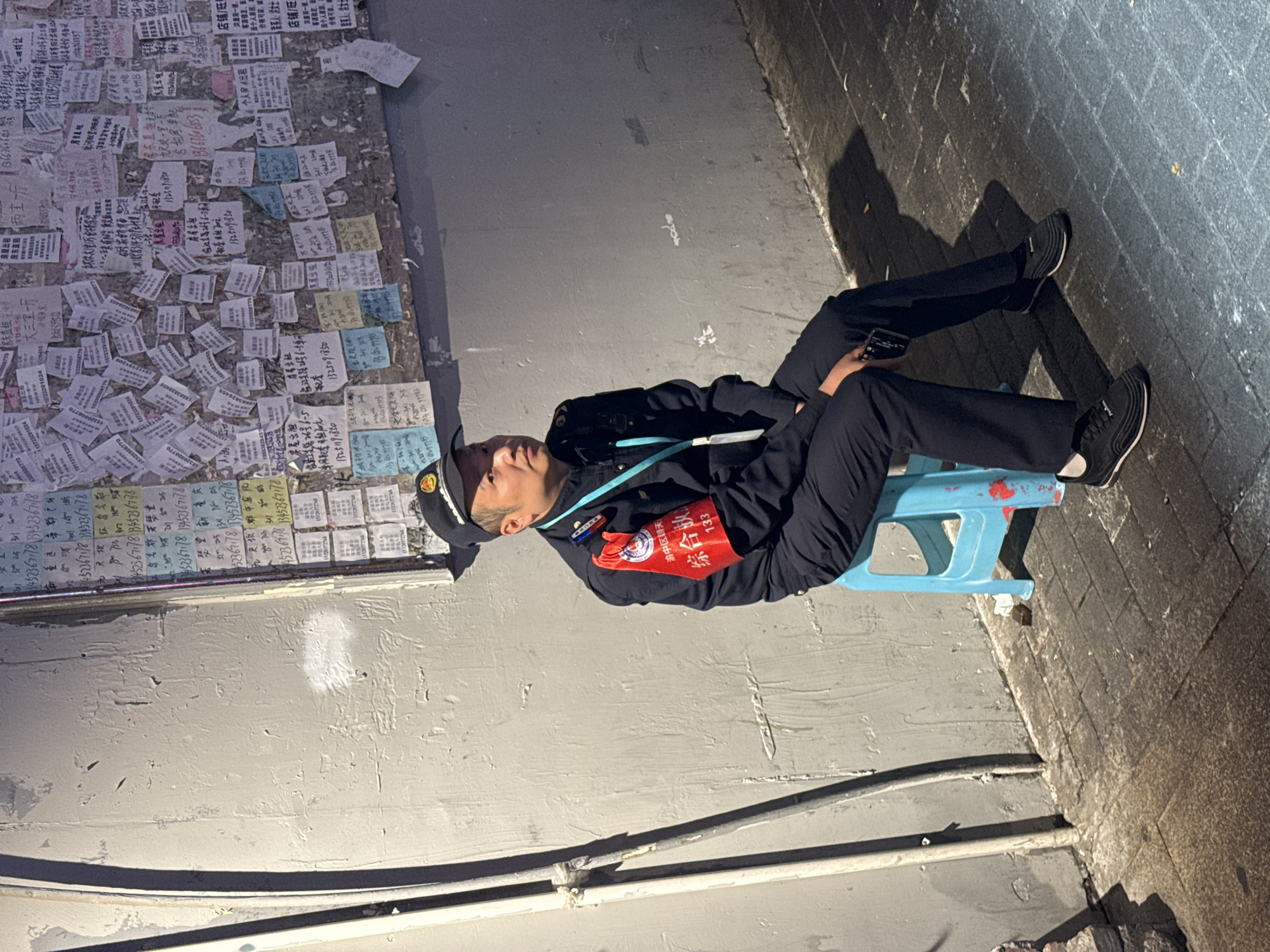 A man in a dark uniform and cap sits on a blue plastic stool in an alley. He is holding a mobile phone. A red armband is on his left arm. The text "渝中区朝天, 综合执 133" is on the armband. A wall covered in numerous advertisements and notices is directly behind him. The notices contain Chinese characters and telephone numbers. He is sitting on the border between the wall with the signs and a rough brick wall, which is on the side.