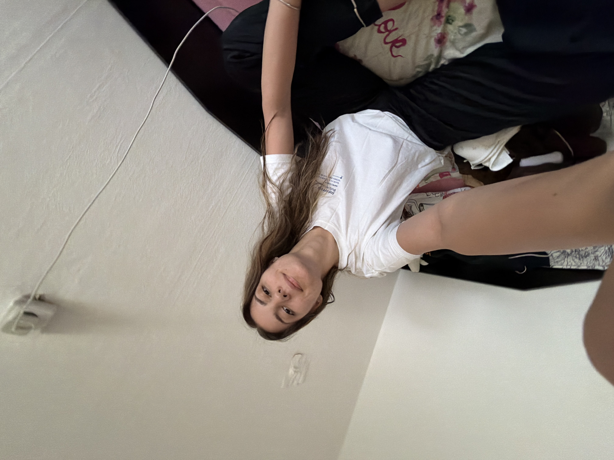 A young woman is in a bedroom, likely indoors, on an upper level of a residence in Sofia, Bulgaria. She is dressed in a white T-shirt and dark pants, seated on what appears to be a bed with a pillow that has the word "love" written on it. An electrical cord is visible coming from the ceiling. The scene is rotated; the viewpoint is upwards from a lying position.