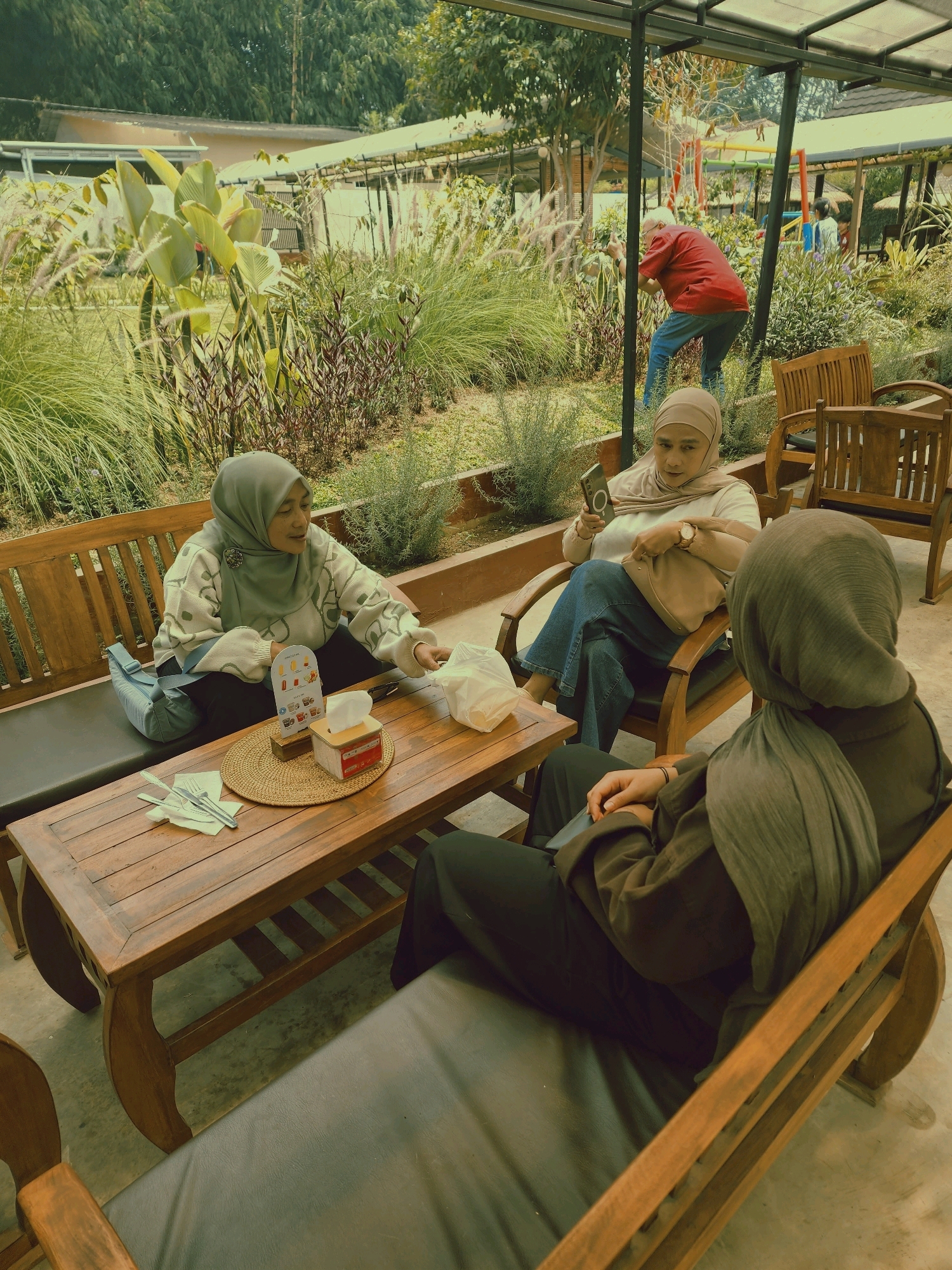 In Langensari, Indonesia, three individuals are seated at a wooden table outdoors. Two women are seated at the table on a wooden bench, facing each other, while a third person is seated on a wooden chair to the right, facing away from the camera. The woman on the left is wearing a grey hijab and a patterned sweater, and is holding a white plastic bag. The woman in the middle is wearing a tan hijab and a white shirt, and is looking at her phone. The person on the right is wearing a grey hijab and a dark brown shirt, and is looking down. On the table, there is a woven placemat, a tissue box, cutlery on a napkin, and a small display stand. The setting appears to be a cafe or restaurant with lush greenery in the background, including tall grasses and plants. A man in a red shirt and blue jeans is visible in the distance, taking a photo of the plants.