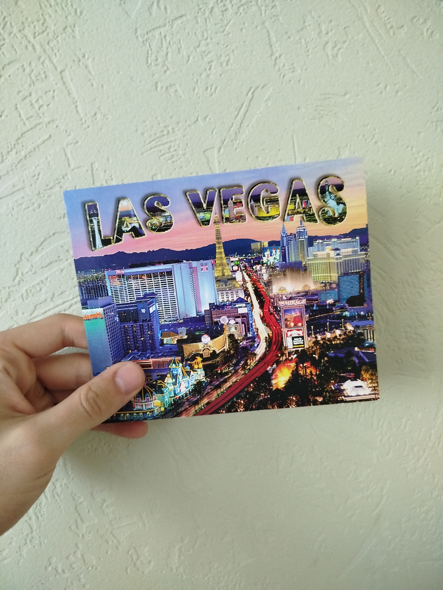 A hand holds a postcard of Las Vegas. The postcard features a cityscape of Las Vegas at dusk, with prominent hotels and casinos visible along the Strip. The words "LAS VEGAS" are displayed in large, decorated letters across the top of the postcard. The background is a textured, light-colored wall.