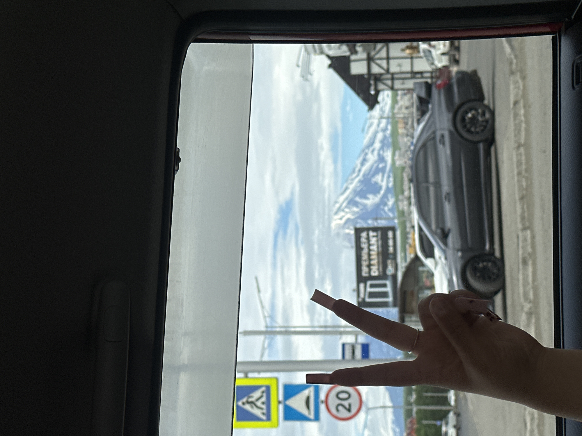 A hand with long, pink, decorated nails is held up in front of a car window, forming a peace sign. Outside the window, snow-capped mountains are visible under a cloudy sky. A dark gray car with a spoiler is parked on the street, and several traffic signs, including a pedestrian crossing sign, a speed bump warning sign, and a 20 km/h speed limit sign, are also visible. A billboard that reads "ПРЕМЬЕРА DIAMANT" is also present. The scene appears to be in the city of Petropavlovsk-Kamchatsky, Russia, based on the combination of elements such as the mountains, architecture, and Cyrillic signage.