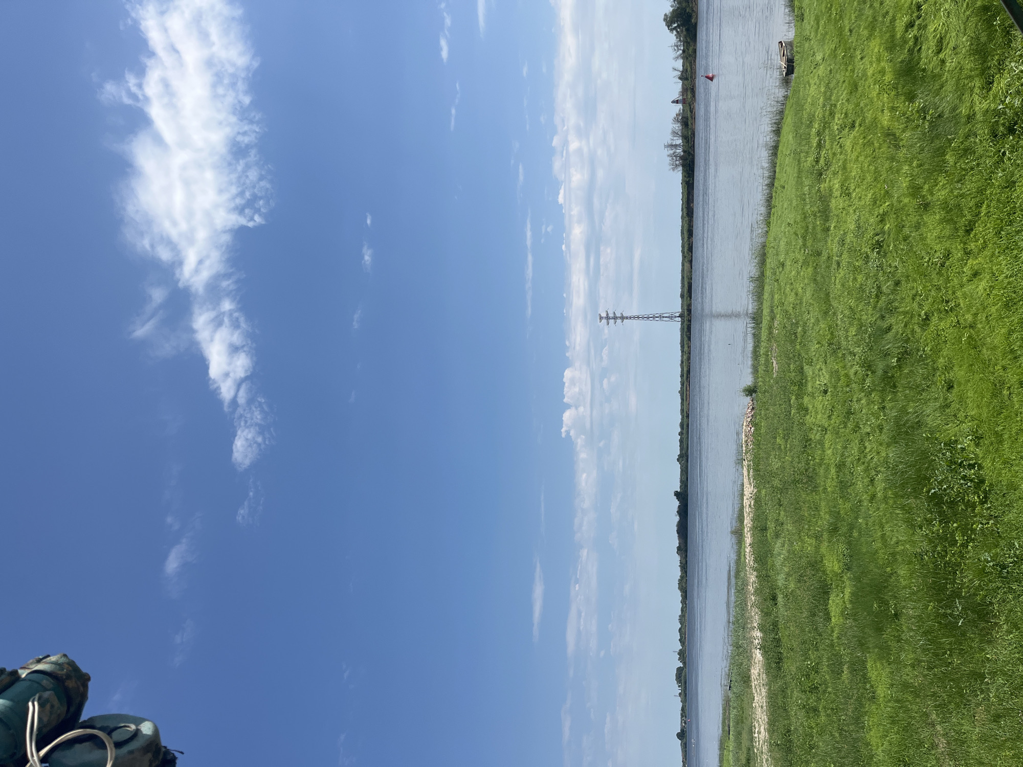 The image depicts a wide, calm body of water, likely a river or estuary, under a bright blue sky with scattered white clouds. The foreground is dominated by a vibrant green grassy field. In the mid-ground, the water stretches across the frame, with a distant, tree-lined shore visible. A prominent, tall metal lattice pylon stands on the far shore. Near the pylon, a red and white lighthouse-like structure is discernible. A solitary red buoy floats on the water in the mid-right of the image. A small, weathered boat rests on the grassy bank in the lower right corner. The top left corner of the image shows a portion of what appears to be rigging or equipment, possibly from a boat or a dock structure. The scene is peaceful and suggests an outdoor recreational or industrial area.