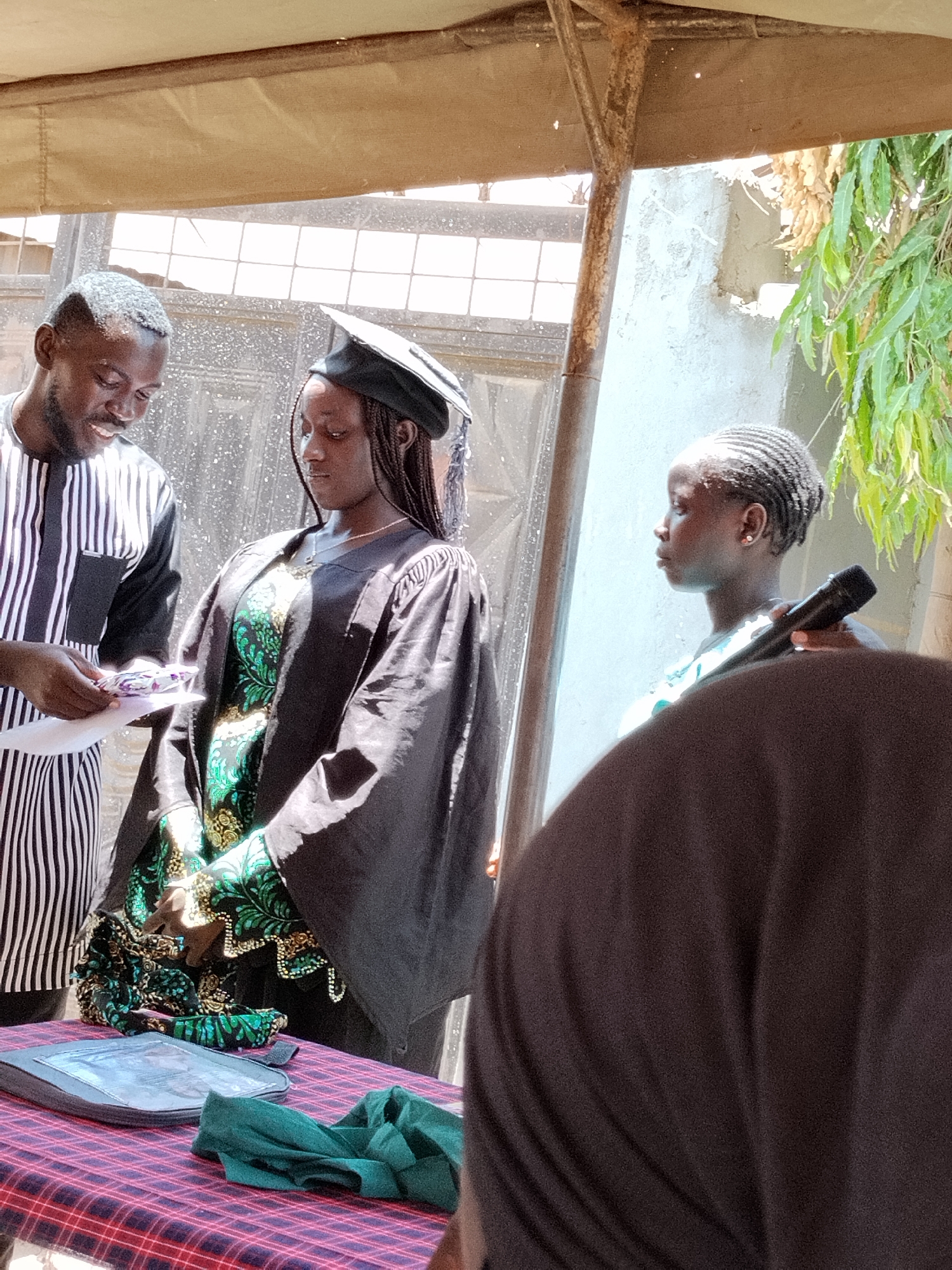 Three individuals are present outdoors under a makeshift canopy. A man wearing a vertically striped black and white shirt is handing papers to a woman in a graduation gown and cap. The woman wears a green patterned dress under the gown. A girl stands to the right, holding a microphone. A table covered with a red and blue checkered cloth holds a case and a green cloth. The setting is Sabon Tasha, Nigeria, evidenced by architectural style and vegetation.
