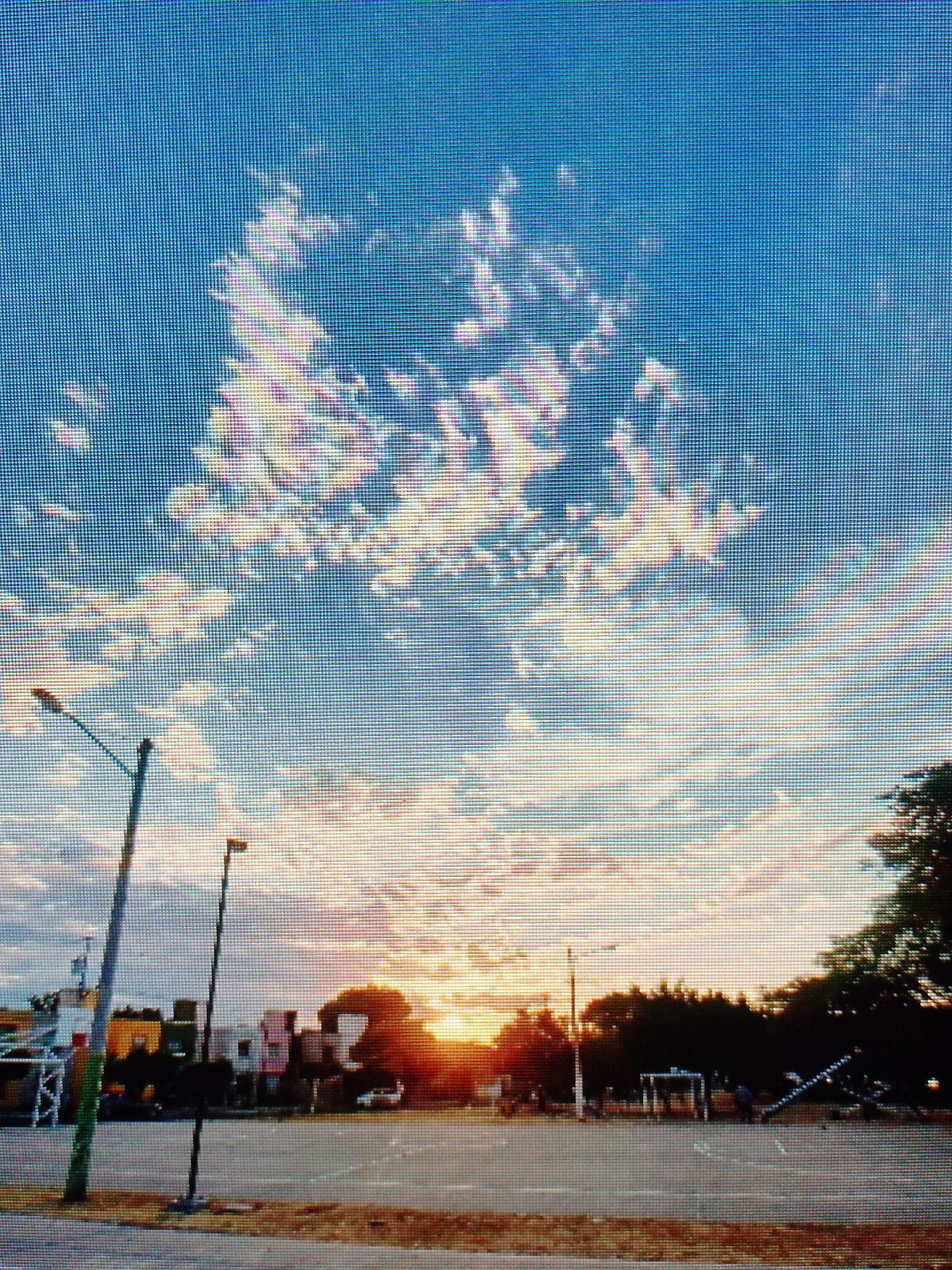 The image shows a street scene at sunset. The sky is a mix of blue and orange hues with scattered clouds. A parking lot occupies the foreground, with streetlights, trees, and buildings visible in the background. A white car appears parked behind the silhouetted trees. In the right corner there is a treeline. No people are clearly visible. The location is given as Unknown, Unknown.