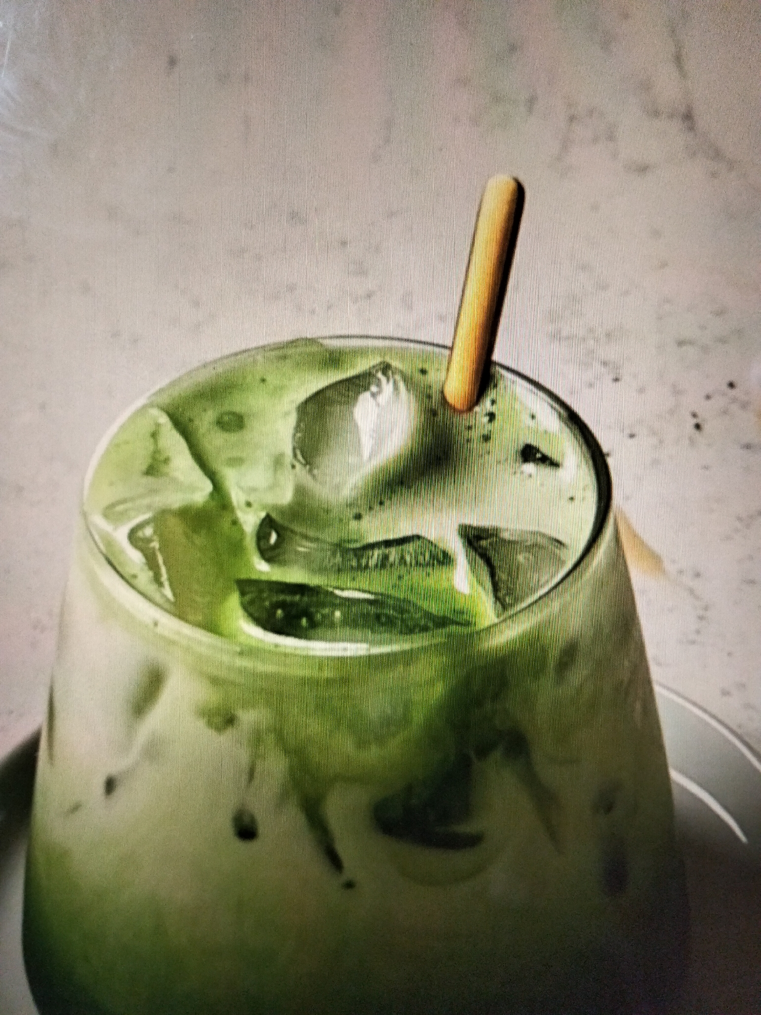 A close-up, top-down view shows a glass of matcha latte with ice and a wooden stirrer. The drink is vibrant green with streaks of lighter green from the milk. The background is a light gray, textured surface. The image is well-lit, highlighting the condensation on the glass and the texture of the drink. The location in Payagaboh, Indonesia is not discernable from the image.