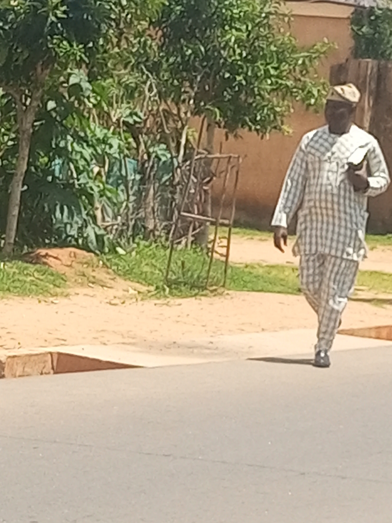 A man wearing a white and grey checkered traditional outfit and a matching cap walks on a paved road in Tempure, Nigeria. He is carrying a dark-colored object in his right hand. The man is walking from right to left in the frame. Behind him, there is a patch of green grass and dry brown earth, with some vegetation and trees. A dark metal structure, possibly a fence or a stand, is visible behind him. The background shows a brown wall and more foliage. The sun is casting a shadow to the left of the man. The image appears to be taken during the daytime.