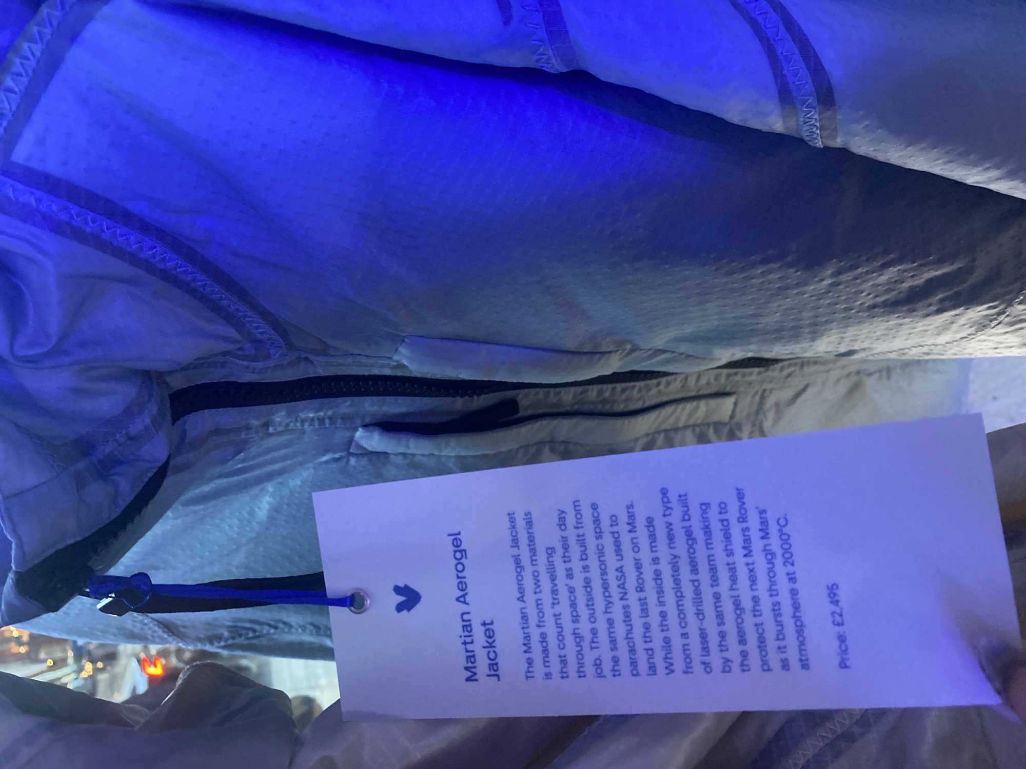The image shows a close-up of a "Martian Aerogel Jacket," with a tag attached that describes the jacket's construction using materials related to space travel. The tag details that the outer layer is made from the same hypersonic space parachutes NASA used to land the last Rover on Mars. The inside of the jacket is made from a laser-drilled aerogel built by the same team who are working on the heat shield to protect the next Mars Rover as it enters the Martian atmosphere. The price of the jacket is listed as £2,495. The environment appears indoors, with the jacket partially visible and blue lighting present.