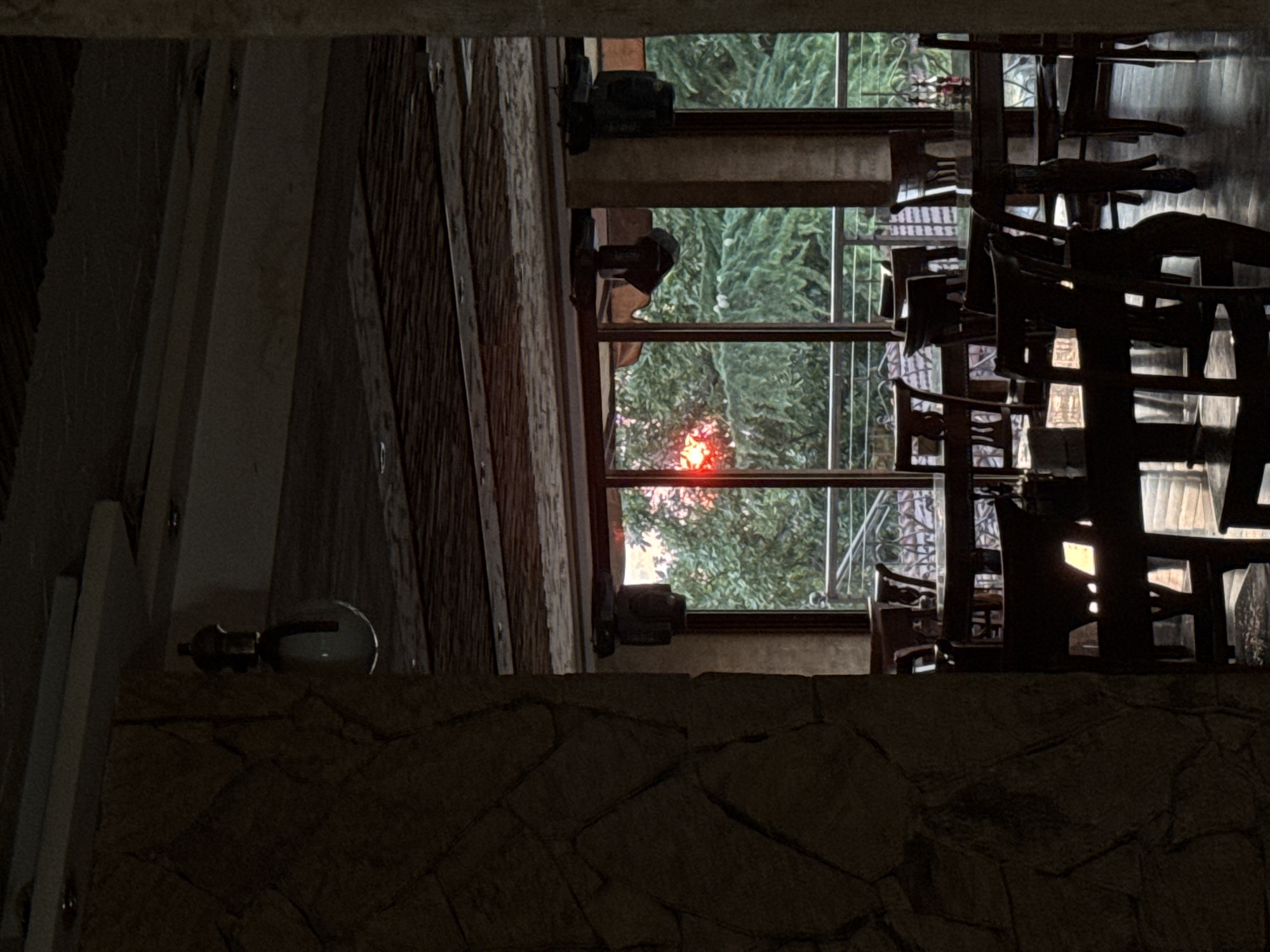 The image depicts the interior of a restaurant in Sredets, Bulgaria, with a view of trees and a sunset through a large window. The restaurant appears to be empty of patrons. The foreground is dominated by dark wooden tables and chairs, with some light filtering through the gaps in the chairs. The walls are made of stone in the foreground and a lighter material further back. The ceiling is paneled with wood and features exposed beams. Several spotlights are visible on the ceiling. The window reveals lush green trees, suggesting an outdoor seating area or balcony beyond. A bright red sun is visible setting through the trees, casting a warm glow. The overall atmosphere is dimly lit and quiet, possibly indicating the end of the day or early evening.