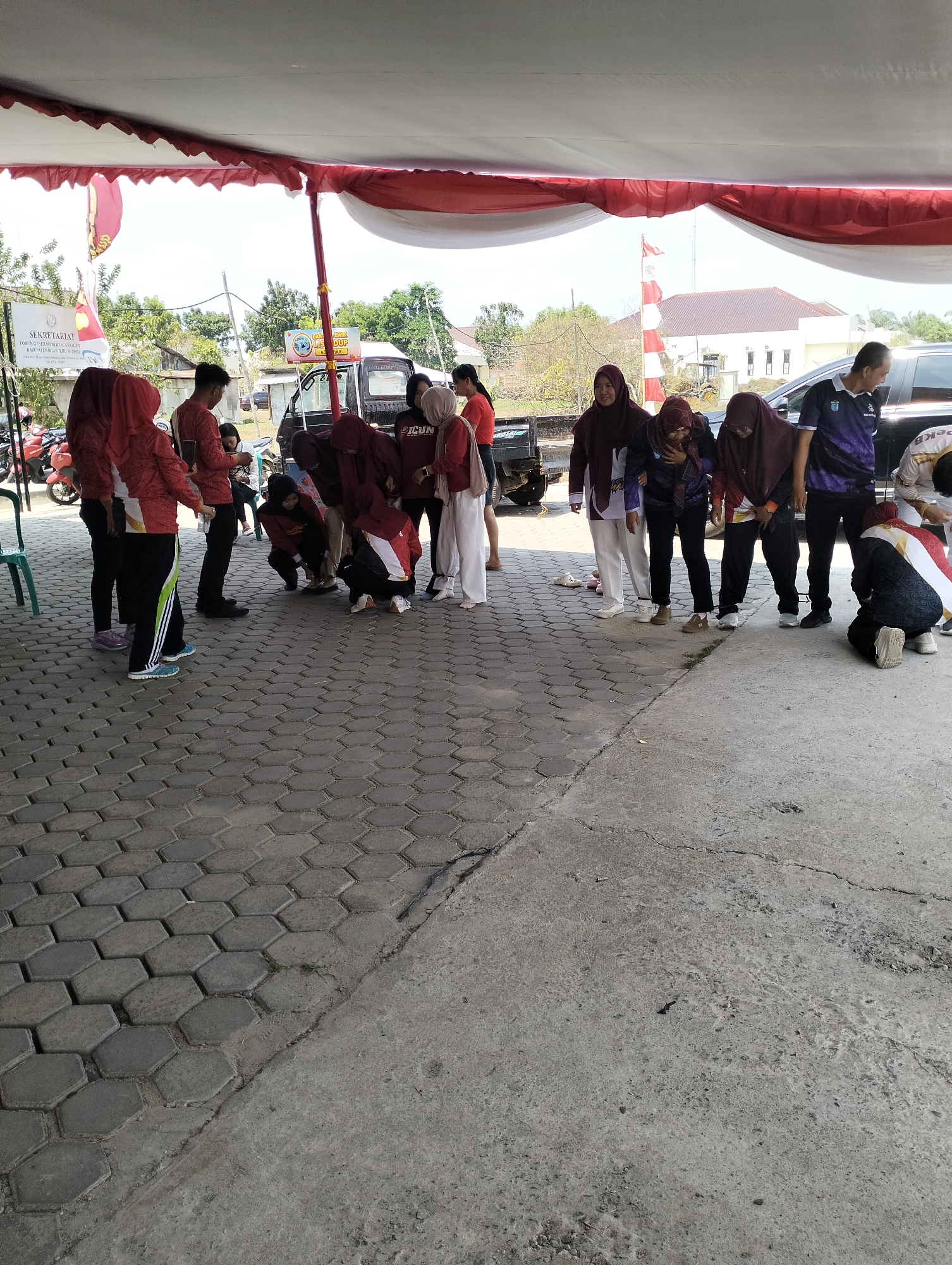 In the city of Tanjung Sejaro, Indonesia, a group of approximately twelve people, mostly young adults, are gathered under a white and red tent. The ground is paved with hexagonal grey bricks and transitions to a cracked concrete surface. The people are dressed in casual attire, with several individuals wearing red or maroon shirts or hijabs, and others in darker colored clothing. Some are engaged in an activity that involves looking down at the ground, with some in crouching or kneeling positions. One individual is wearing a dark blue and purple shirt with a logo. In the background, there are parked vehicles, including a dark-colored van and a truck, as well as buildings and trees. A flag pole with a flag is visible in the middle ground. The lighting suggests it is daytime.