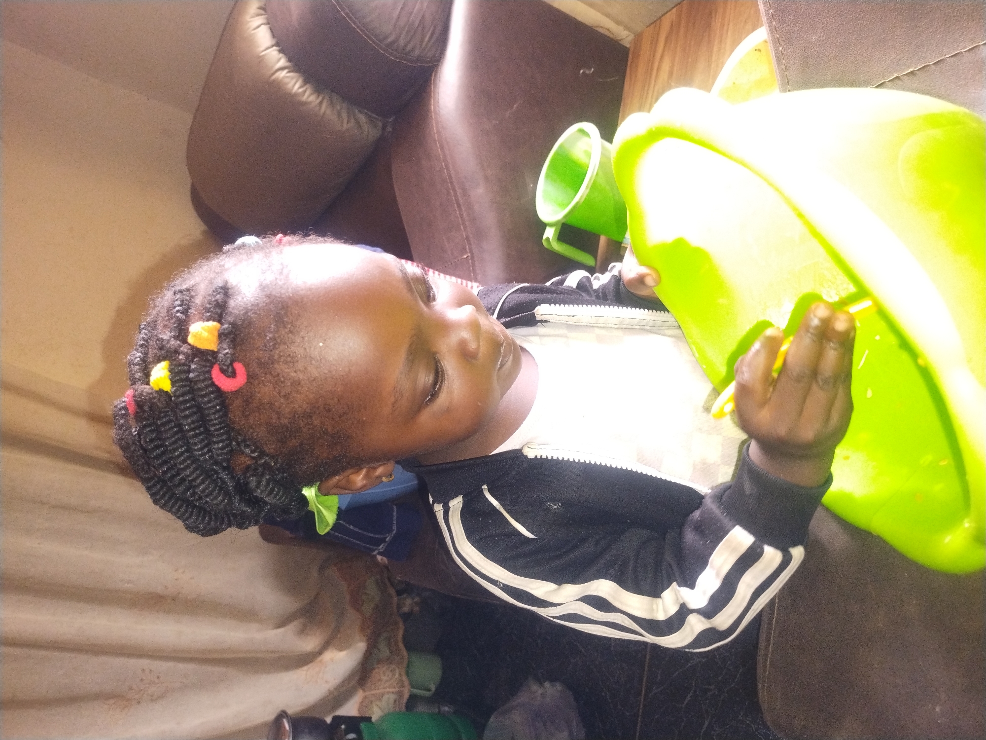A close-up, eye-level shot captures a young Black child wearing a black and white striped jacket over a white t-shirt. The child's hair is styled in dark braids, adorned with small colorful hair clips in red, yellow, and orange. The child is looking down and to the right, holding a large, bright green plastic basin with both hands. One hand is visible holding the edge of the basin, while the other rests inside it, fingers extended. To the right and slightly behind the child, a smaller green plastic cup with a handle is visible. The background features a textured, light-colored curtain on the left, and a brown upholstered piece of furniture, possibly a couch or chair, on the right. The floor appears to be dark and tiled. The lighting is bright, causing some highlights on the child's face and the plastic basin.