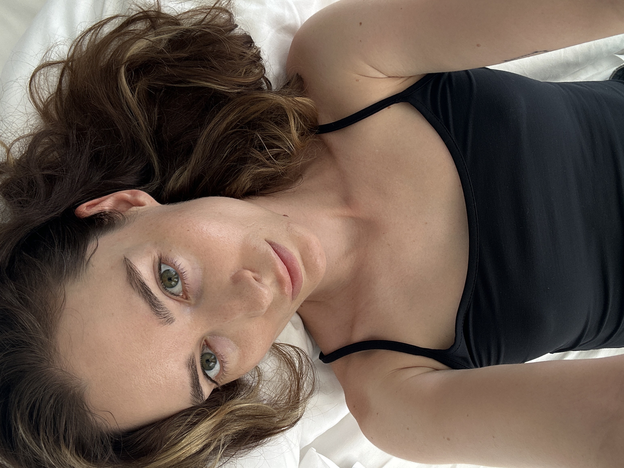 A woman is lying on a white sheet, possibly a bed, wearing a black tank top. She has brown hair with blonde highlights and green eyes. The photo is taken from a high-angle perspective, focusing on her face and upper body. The location, Choeng Thale, Thailand, is not evident from the image.
