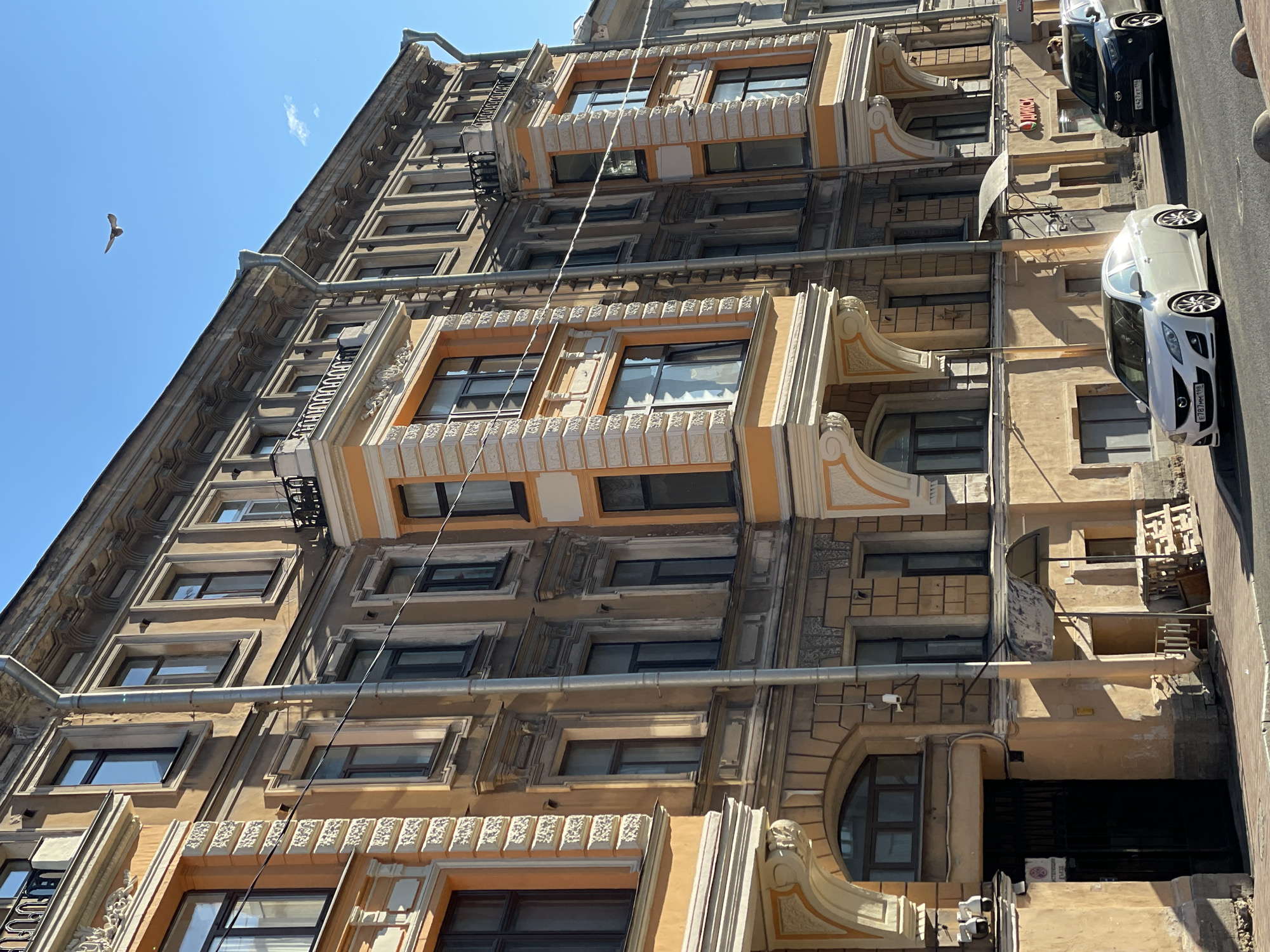 A tall, ornate building is shown in the photograph. The building's facade features a mix of architectural styles, including decorative window frames, projecting balconies, and a series of ornate ledges and moldings. The building's color palette is dominated by shades of tan and orange. A gray metal pipe runs vertically down the building.
Two cars, one black and one white, are parked on the street next to the building.
In the sky, a single bird is visible.
The architecture suggests the image was taken in Saint Petersburg, Russia.