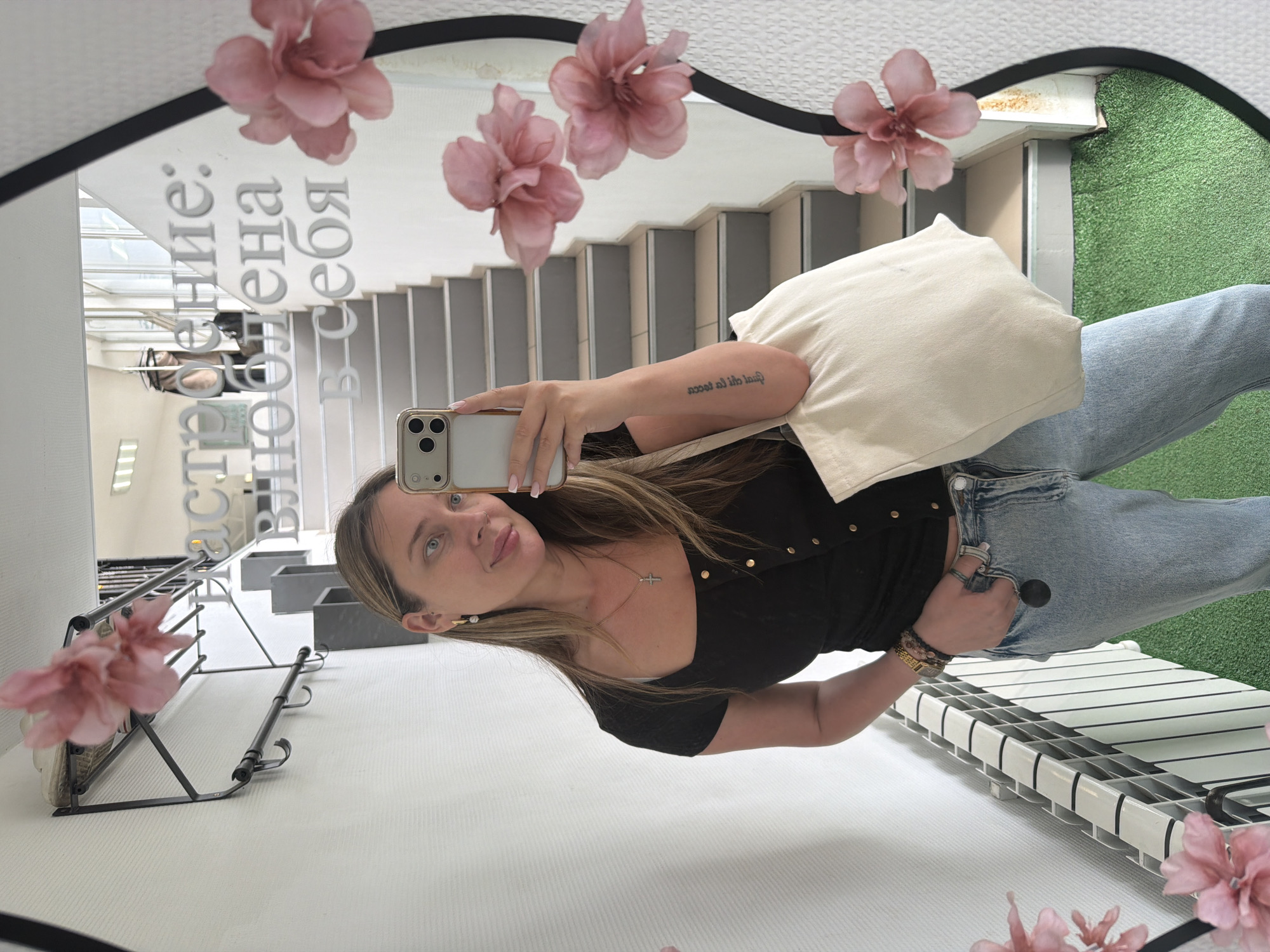 This media file captures a woman taking a mirror selfie, presented with a 90-degree clockwise rotation. The subject is a woman with long, light brown hair and blue eyes, smiling subtly as she looks into the camera of her light-colored iPhone, which she holds with her right hand. She is wearing a black top with gold buttons, light blue jeans, and a white tote bag is slung over her left shoulder. Accessories include a cross necklace, pearl drop earrings, rings on her left hand, and a multi-strand bracelet on her left wrist. A tattoo on her right forearm reads "non alii te ipsum," a Latin phrase meaning "no other than yourself" or "not others, but yourself."
The selfie is taken in an oval-shaped mirror with a black frame, adorned with several pink artificial flowers. The reflection in the mirror reveals an interior setting. Behind the woman, a flight of grey stairs is visible, and to the right of the stairs, there's a patch of green artificial turf. A white slatted shelf or rack is seen below the turf. To the left side of the reflection, another dark metal rack, possibly a shoe or coat rack, is present.
Text is visible on the mirror itself, written in Russian: "Настроение: влюблена в себя," which translates to "Mood: in love with myself." The scene appears well-lit, suggesting daytime. The location context for this image is Chaikovsky, Russia.