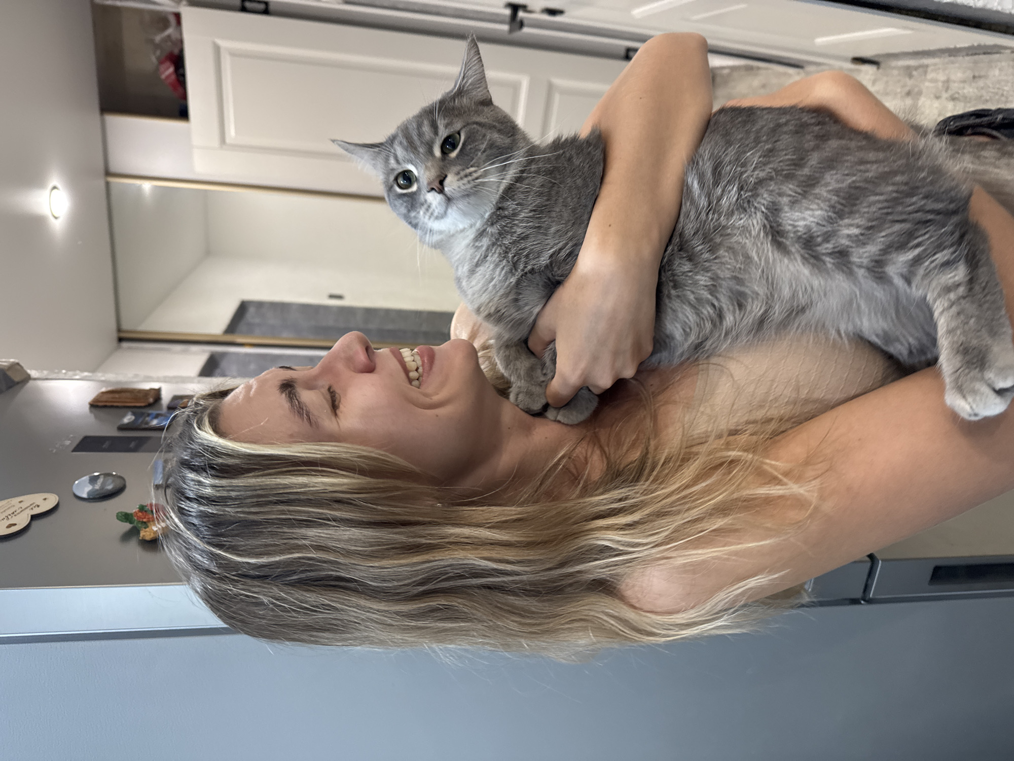 The image is rotated 90 degrees clockwise, depicting a person holding a cat indoors. The subject is a woman with long, wavy blonde and light brown hair, captured mid-laugh or broad smile, showing her teeth. She is holding a medium-sized gray tabby cat, which is nestled against her chest and shoulder area, embraced by her arms. The cat has distinct green eyes and is looking directly at the camera with an alert and curious expression. The woman's expression conveys happiness and affection towards the pet.
The setting is an indoor environment, likely a home, with elements suggesting a kitchen or living area. In the background, white cabinets are visible, along with a bright round light fixture on a light-colored wall to the left. A darker countertop or surface runs beneath the cabinets. To the right, a metallic surface with several small magnets, including one shaped like a turtle, suggests the presence of a refrigerator. The lighting appears to be artificial, originating from the visible light fixture. No discernible text is visible in the image.