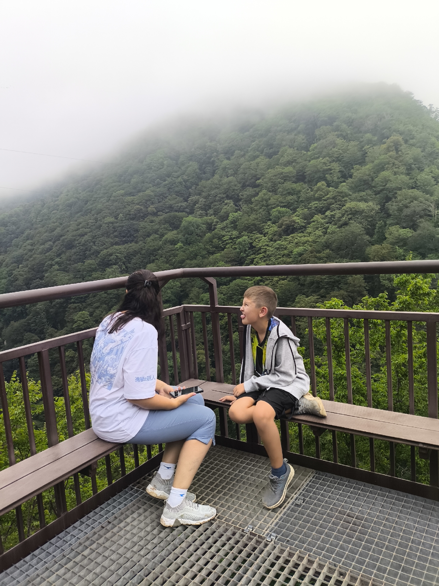 Two people are sitting on a wooden bench on a metal grate platform. The person on the left, wearing a white t-shirt and blue capri pants, is a female with long dark hair and is holding a phone. The person on the right, a young boy wearing a grey hooded jacket and black shorts, is sticking out his tongue. They are facing each other, and appear to be in conversation. The background is a lush green mountain covered in trees, with fog obscuring the top of the mountain. The location is the Gaghma Nogha scenic viewpoint in Gaghma Nogha, Georgia.