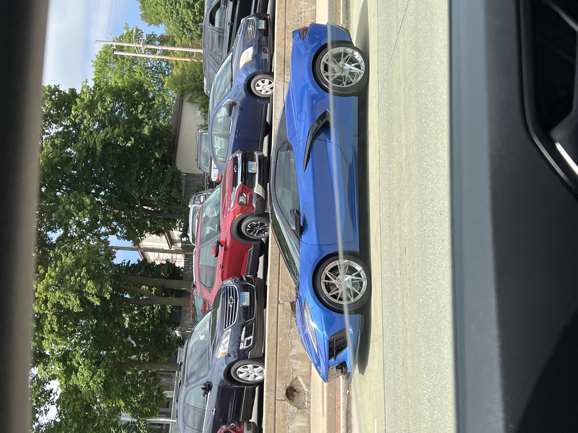 The image captures a street scene with several parked vehicles along a curb. The most prominent vehicle is a bright blue sports car, likely a Chevrolet Corvette, with chrome wheels. It is parked next to a navy sedan, a red SUV, and a black SUV. Other vehicles are partially visible in the background. The street is paved and the curb appears to be concrete. Trees are visible on the opposite side of the street. The photo seems to be taken from inside another vehicle, as part of the interior is in the foreground. The location is a residential or commercial area with street parking.
