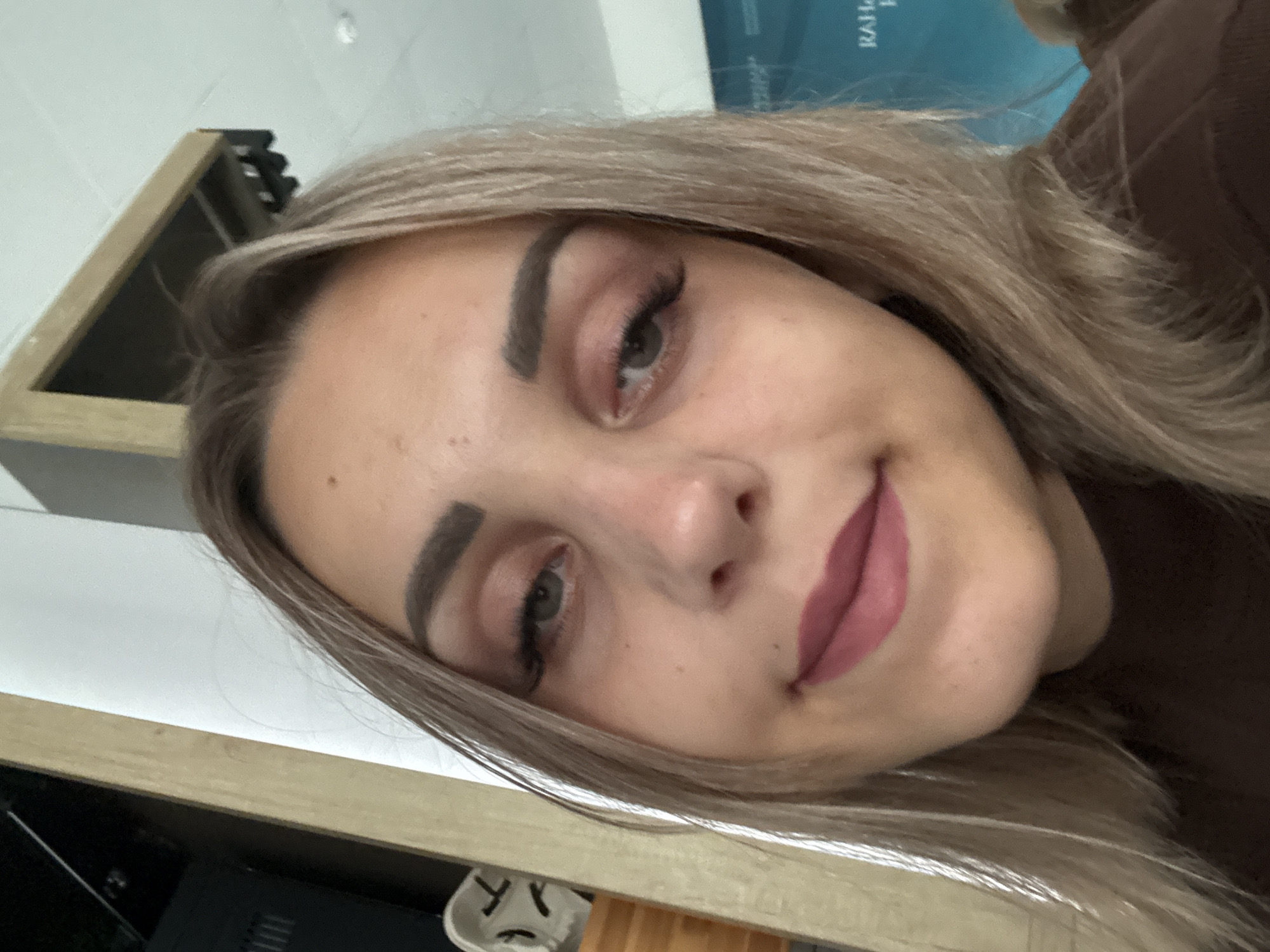 A young woman with blonde hair and makeup takes a selfie. She is looking directly at the camera, with her head slightly tilted. She is wearing a brown shirt. The background is blurred and appears to be indoors, with a wooden structure and a white wall visible. There is also a blue sign in the background with some text on it.