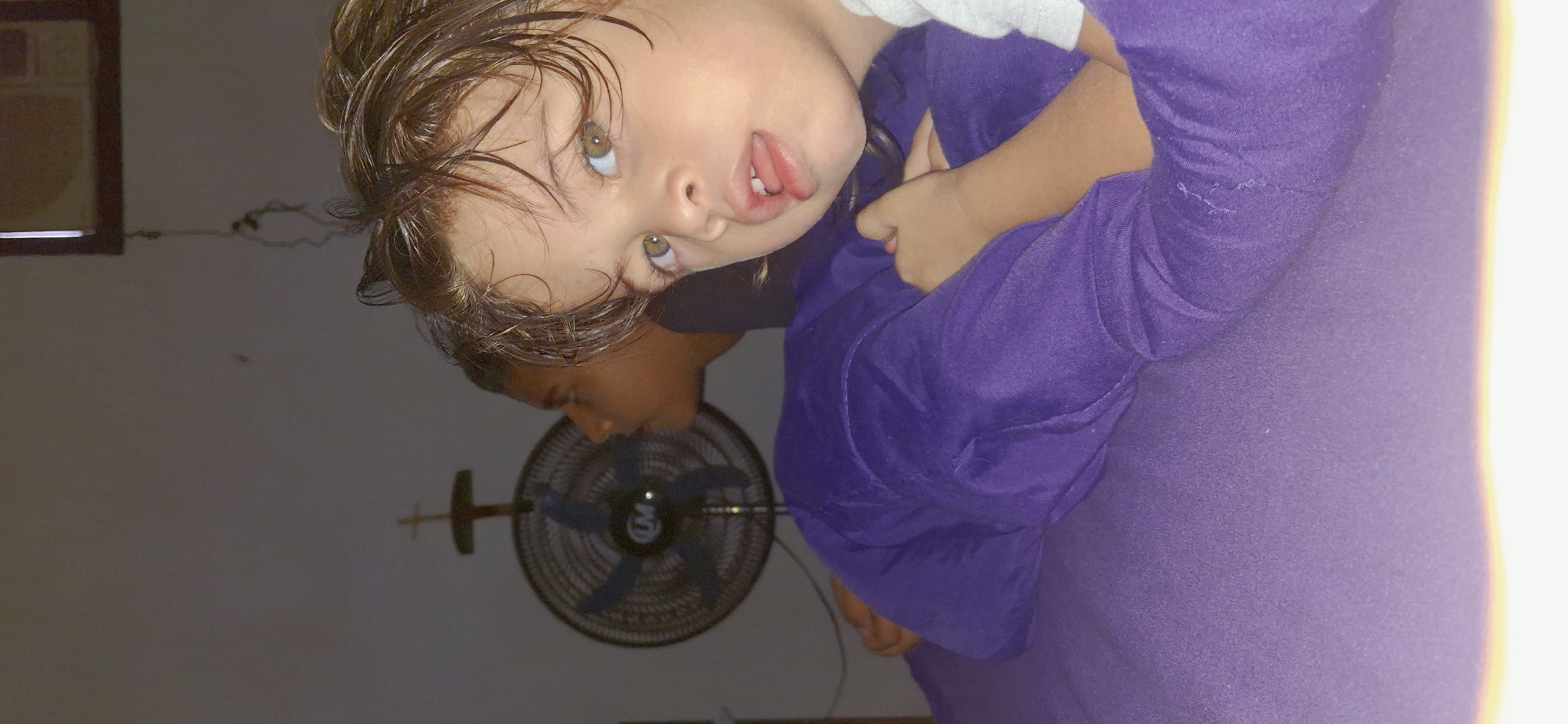In Vallecito, Venezuela, an indoor scene shows two young children on a purple surface, possibly a bed or couch. The child in the foreground, facing the camera, has light brown hair and green eyes. Their mouth is slightly open, revealing their tongue. They are wearing a light-colored shirt, and their left arm is resting on the purple surface. Behind them, another child, possibly a boy, is visible from the side, looking to the left. A black oscillating fan with blue blades is mounted on the wall behind the second child, with a small cross-shaped ornament above it. The wall appears to be painted a neutral color.