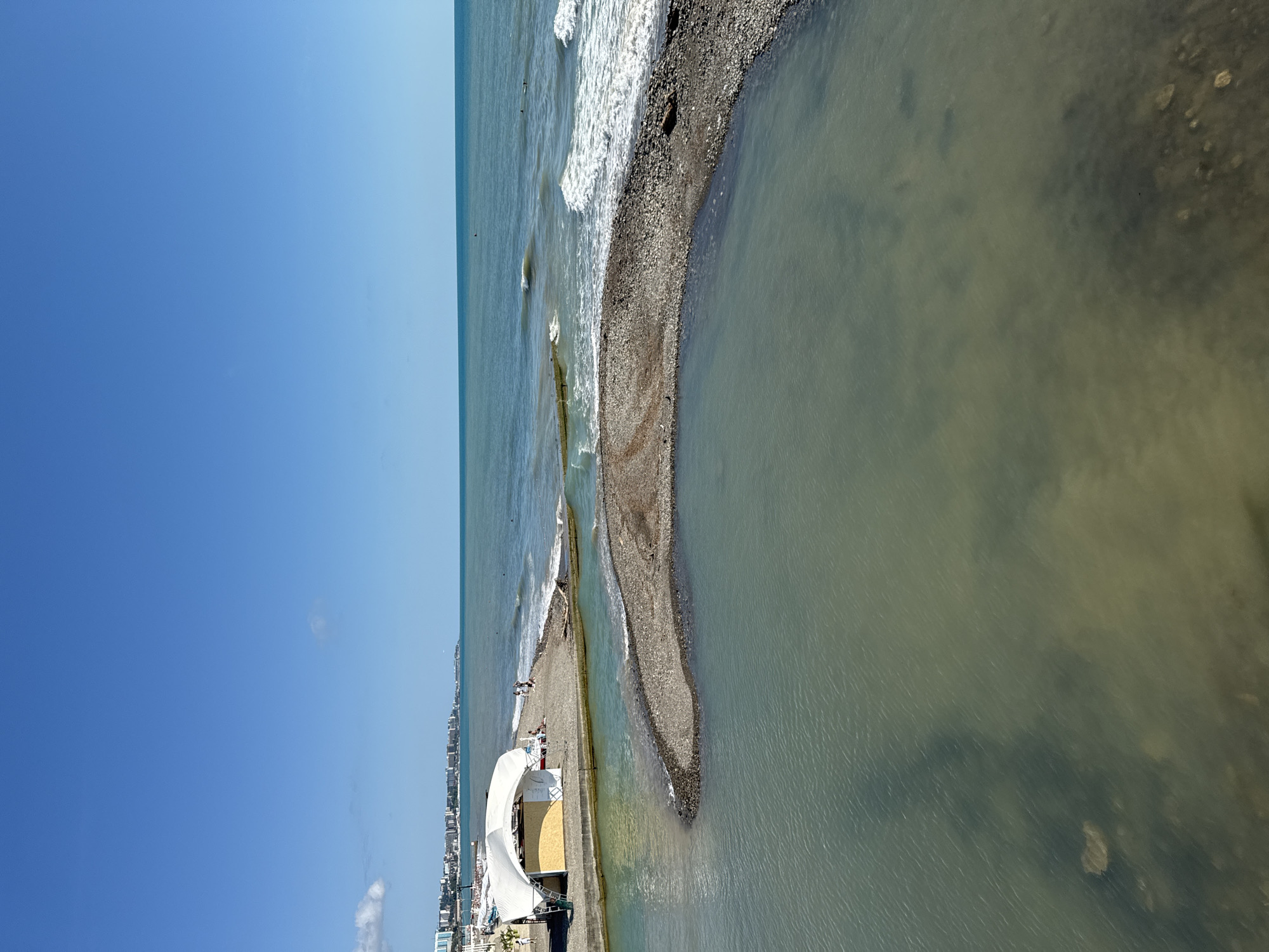 A wide, high-angle shot captures a coastal scene with a clear blue sky above and a body of water in the foreground. The water in the foreground is calm and appears somewhat murky, with visible rocks and debris at the bottom. A narrow, pebble-strewn strip of land juts out into this body of water, separating it from the ocean.
Beyond this strip of land, the ocean is visible with moderate waves breaking on a pebble beach. A white, tent-like structure with a curved roof is situated on the beach, with a few people visible around it. The beach itself has a few lounge chairs and umbrellas. Further down the coastline, a city skyline with numerous buildings is visible, stretching along the shore. The horizon line is clearly defined between the sea and the sky.
This image depicts a section of the Sochi coastline in Russia, likely near a river mouth or estuary where freshwater meets the sea. The presence of the tent structure, beach amenities, and the city in the background are indicative of a populated resort area.