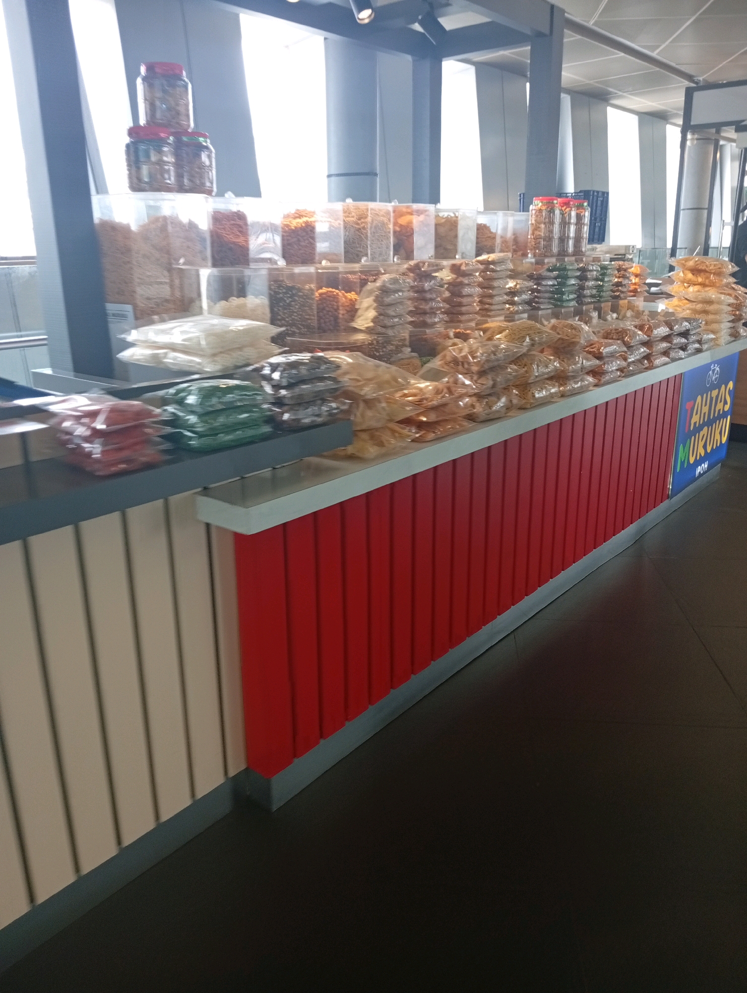 The image shows a food stall in a modern building, likely an airport or a shopping mall. The stall is filled with various packaged snacks and treats, displayed in clear plastic bags and jars. The stall's facade is a mix of white and red vertical panels, with a grey countertop. A blue sign with white text reading "TAHTAS MURUKU IPOH" is visible on the right side of the stall. Natural light streams in from large windows in the background, illuminating the interior of the building which features structural beams and ceiling elements. The floor is dark and reflective. There are no people visible in the image. This scene is likely located in Kuala Lumpur International Airport (KLIA) as "Ipoh" in the sign suggests a product from Ipoh, a city in Malaysia, and such food stalls are commonly found in Malaysian airports.