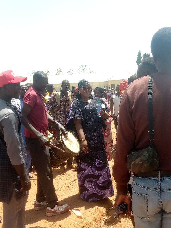 The image depicts an outdoor gathering under bright sunlight in Kagoma, Nigeria. The ground is reddish-brown dirt. In the foreground, a man wearing a red shirt, dark pants, and white sneakers plays a djembe-style drum with a stick. To his right, a woman in a long, dark purple patterned dress, dark headwrap, and large sunglasses smiles, holding a clear plastic water bottle in her left hand and a dark smartphone in her right. Gold-colored bracelets are visible on her left wrist. To the left of the drummer, a man in a red baseball cap and light-colored top is partially visible. The back of another man, wearing a brown long-sleeved shirt, blue jeans, and a camouflage shoulder bag, occupies the right foreground, holding a dark smartphone and keys in his right hand. A crowd of individuals in various attire is visible in the mid-ground and background. A light-colored building with a white roof and some trees are discernible in the distant background. A small, white, elongated object rests on the ground near the drummer's feet.