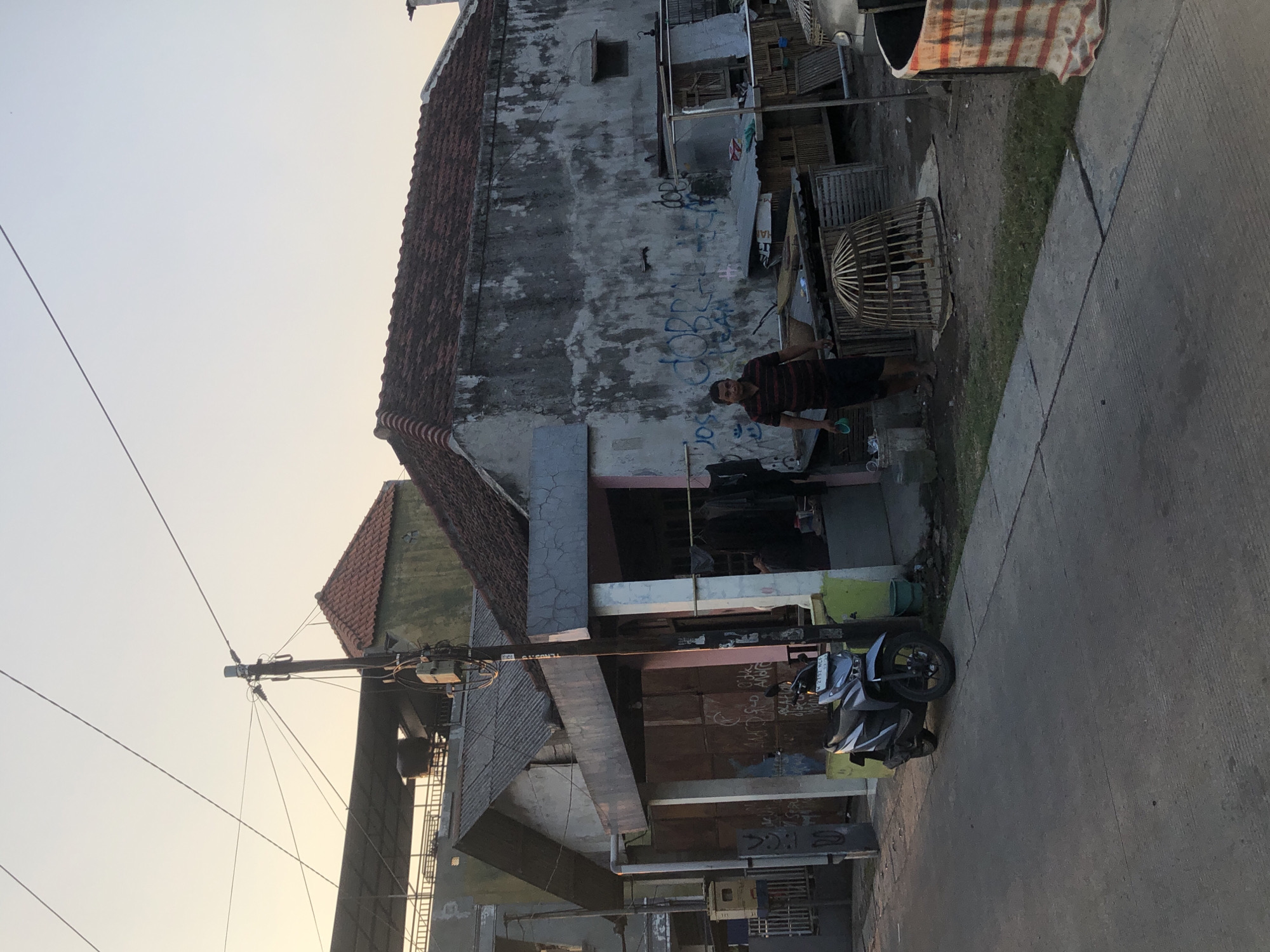 The image depicts a street scene in Tenggulunan, Indonesia, at dusk. A man in a striped shirt and dark shorts stands next to a wooden chicken coop. In the foreground, a gray motorcycle is parked on the concrete road. Adjacent to the road are several buildings with weathered walls and tiled roofs. A utility pole with wires runs vertically through the scene, connecting to the structures. The sky is a pale orange, indicating the setting sun. The environment appears to be a residential or semi-rural area, with a mix of structures and outdoor elements.