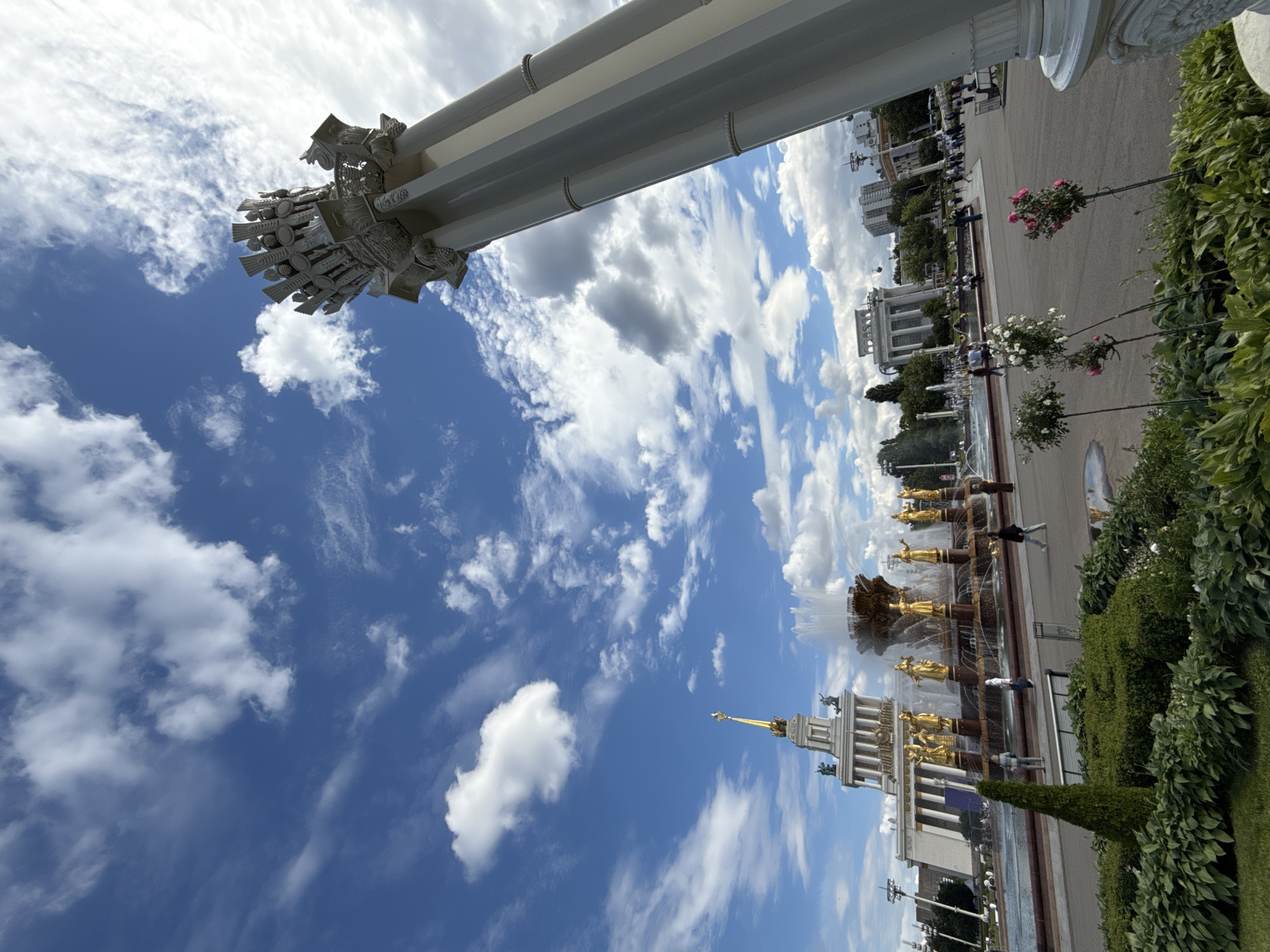 The image depicts a large fountain with golden statues, likely the Friendship of Nations Fountain at VDNKh in Moscow, Russia. The fountain is surrounded by lush green foliage and a paved walkway where several people are visible. In the background, there are grand Soviet-era buildings, characteristic of the VDNKh exhibition center. The sky is a bright blue with scattered white clouds, suggesting a pleasant, sunny day, likely in the late morning or early afternoon. The perspective of the shot is looking upwards, emphasizing the imposing scale of the architectural elements and the vastness of the sky. One of the prominent features in the foreground is a large white column, part of the exhibition center's architecture. The overall scene conveys a sense of grandeur and public space, likely a tourist or recreational area. There is no visible text in the image.