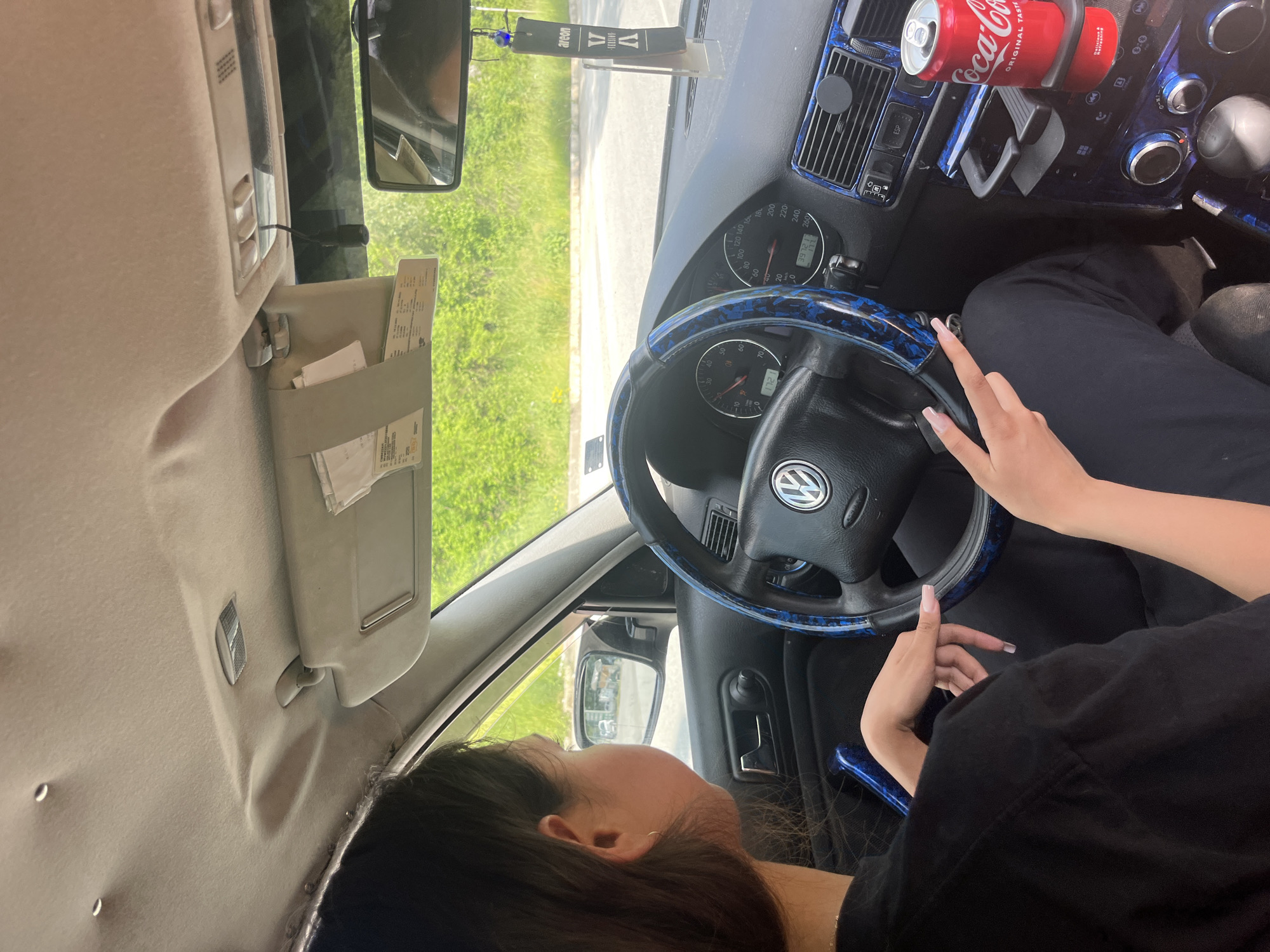 The image depicts the interior of a Volkswagen car, viewed from the passenger's perspective. A young woman, with dark hair and a black shirt, is seated in the driver's seat. Her hands are on the steering wheel, which is adorned with a blue and black patterned cover. The dashboard displays the car's instrumentation, including the speedometer and tachometer. To the right of the steering wheel, in the center console, a can of Coca-Cola is placed in a cup holder. The exterior environment visible through the windows is a sunny day with green foliage, suggesting a rural or suburban setting. The interior of the car shows some wear, with a sagging headliner visible towards the top of the frame. There is an air freshener hanging from the rearview mirror. The car appears to be parked. The location is Vlahovo, Bulgaria.