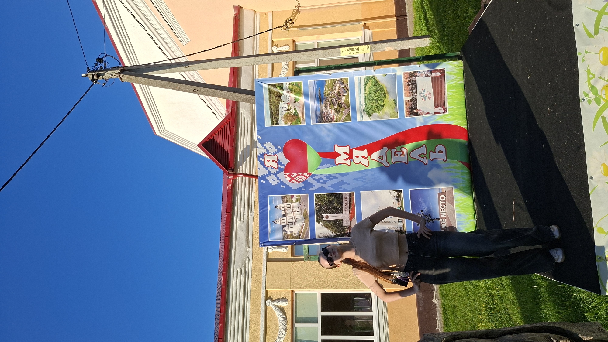 A young woman with long auburn hair, wearing sunglasses, a beige crop top, and dark wide-leg jeans, stands with her hands on her hips in front of a large blue banner. The banner features multiple images of Belarusian landmarks and landscapes, including a prominent red heart with the Belarusian flag. Vertical text in Cyrillic reads "Я МАЁ" (Ya Mayoe), meaning "I am mine" or "My." The scene is set outdoors on a sunny day, with a clear blue sky. Behind the banner, a yellow building with red trim and decorative white elements is visible. A concrete utility pole with electrical wires stands to the right of the banner. The ground in front of the banner is covered with a black mat, and a grassy area is present to the left. The overall impression is a patriotic display, likely at a public event in the city of Myadzyel, Belarus.