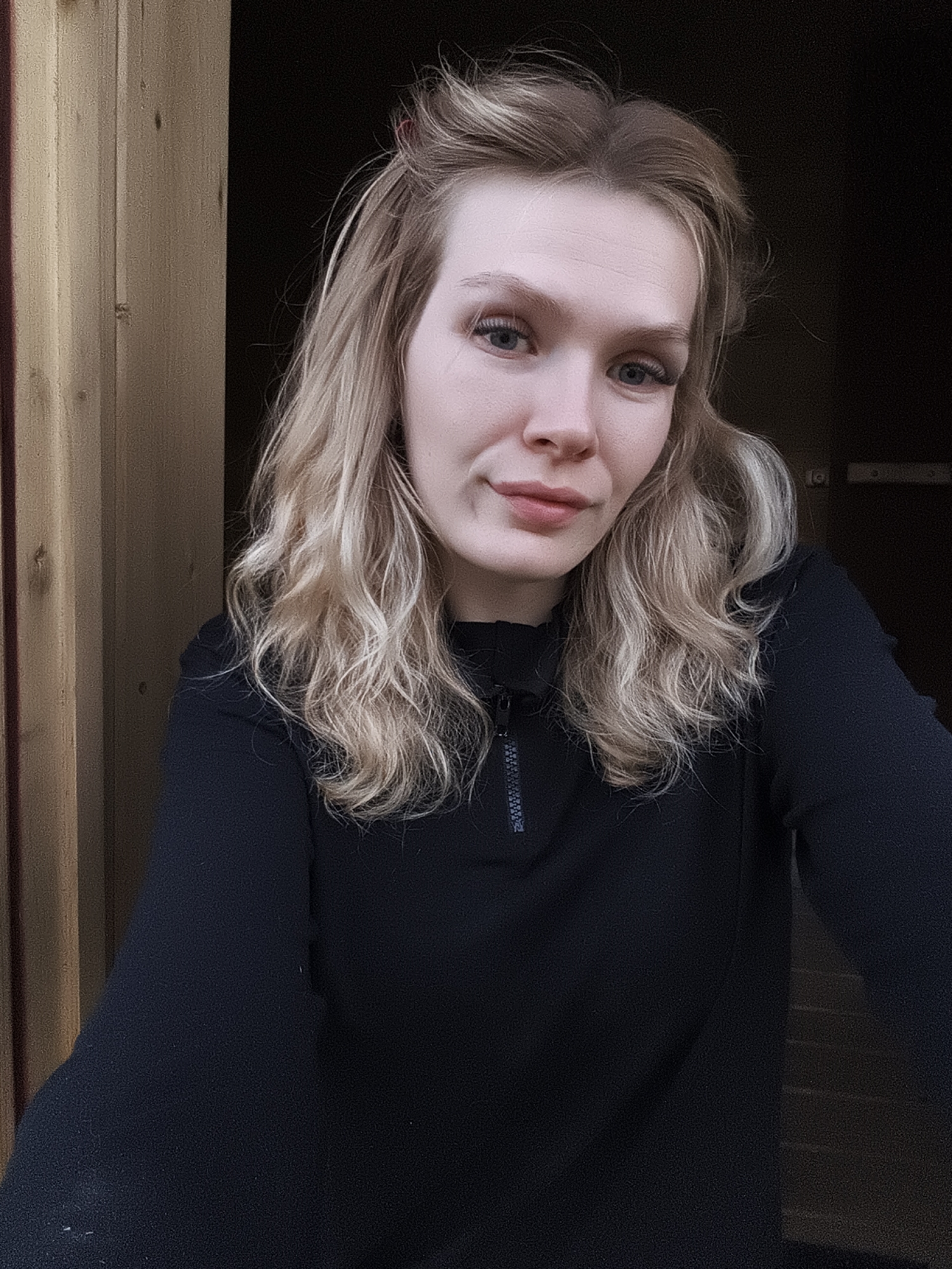 A woman with blonde, shoulder-length wavy hair stands in front of a doorway. She wears a black long-sleeved top with a zipper. The doorway is framed by wooden planks on the left and leads to a darkened interior. The location is Русский Вожой, Russia.