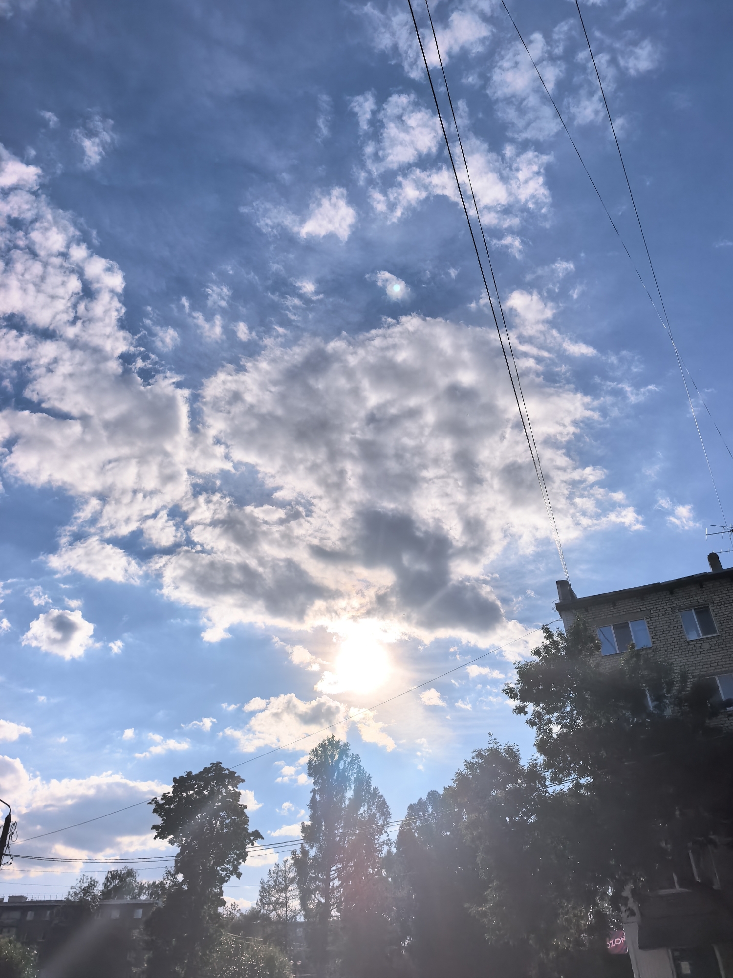 A low-angle shot captures a bright, partly cloudy sky over Sterlitamak, Russia. The sun, partially obscured by clouds, emits strong rays. Overhead power lines stretch diagonally across the frame. In the lower right corner, the top of a brick building with multiple windows is visible, surrounded by the dark silhouettes of trees. The bottom of the image is dominated by dense foliage.