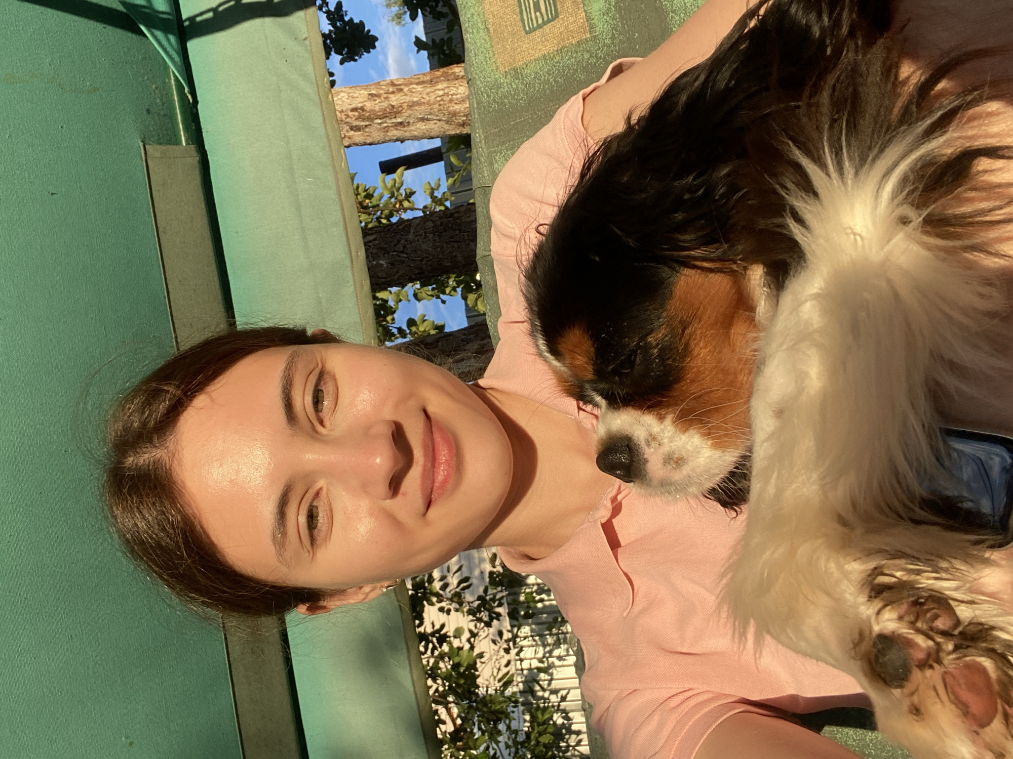 This is a waist-up, selfie-style outdoor photograph of a young Caucasian female with light skin and brown hair, wearing a pink polo shirt. She is holding a Cavalier King Charles Spaniel, which is mostly white with brown and black markings. The dog is being held in her arms, and its face is close to hers, with its nose touching her cheek. The woman is smiling gently at the camera.
The background suggests an outdoor setting, possibly a backyard or park. There is a green canopy overhead, and beyond that, trees and a blue sky with scattered clouds are visible. The woman appears to be sitting on a bench or swing with a patterned cushion. The lighting indicates it is daytime, with the sun casting warm light on the woman's face. There are no discernible landmarks that would indicate a specific location within Votkinsk, Russia.