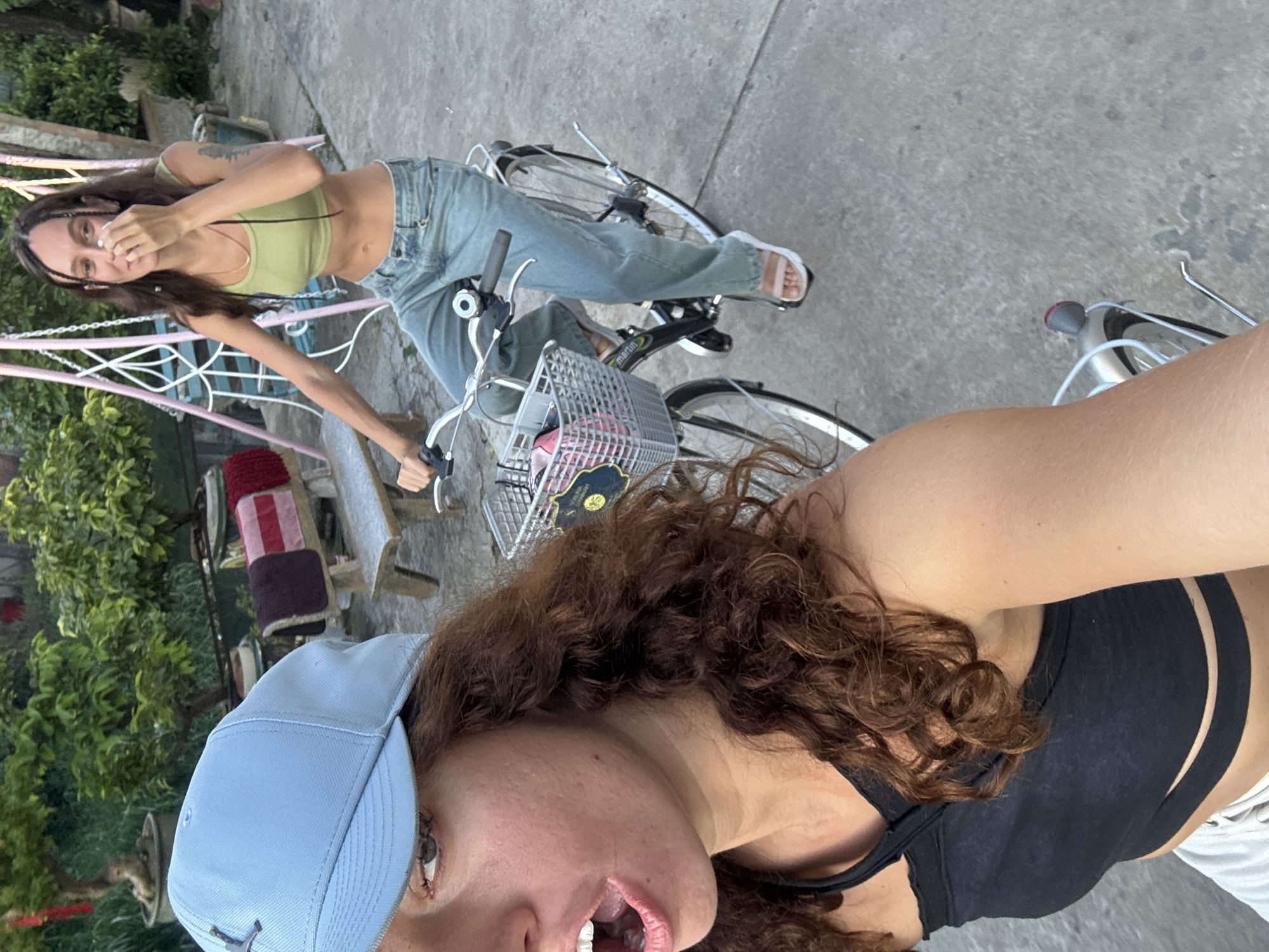 Two women are riding bicycles. The woman in the foreground is wearing a black sports bra and a light blue baseball cap. The woman behind her is wearing a light green crop top and light wash jeans. They are on a paved surface with a building and greenery behind them. The city of Đà Nẵng is not definitively discernible from the provided image.