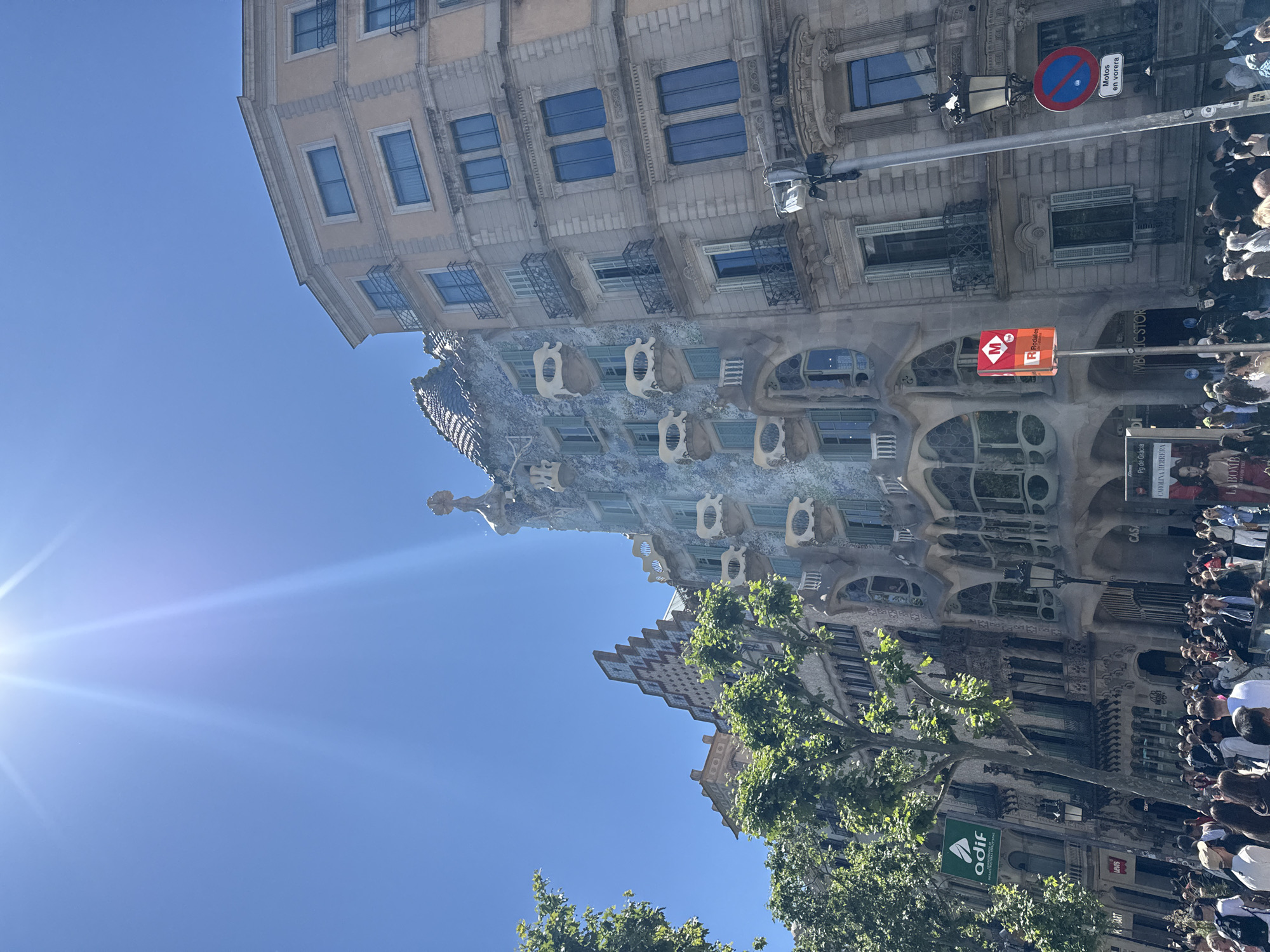The image is an eye-level, slightly tilted view of Casa Batlló, an iconic building in Barcelona, Spain. The building is characterized by its undulating facade, skeletal balconies, and colorful, mosaic-tiled roof. Adjacent to Casa Batlló is a more traditional building, with a simpler facade and rectangular windows. The sky is bright blue, with the sun shining in the upper left corner. A street lamp with a "Motos en Vorera" sign is visible. Several people are on the street, suggesting a tourist location. A tree with green leaves is visible in the lower part of the image, along with a sign that says "ADIF." Various signs are also visible on the building.
