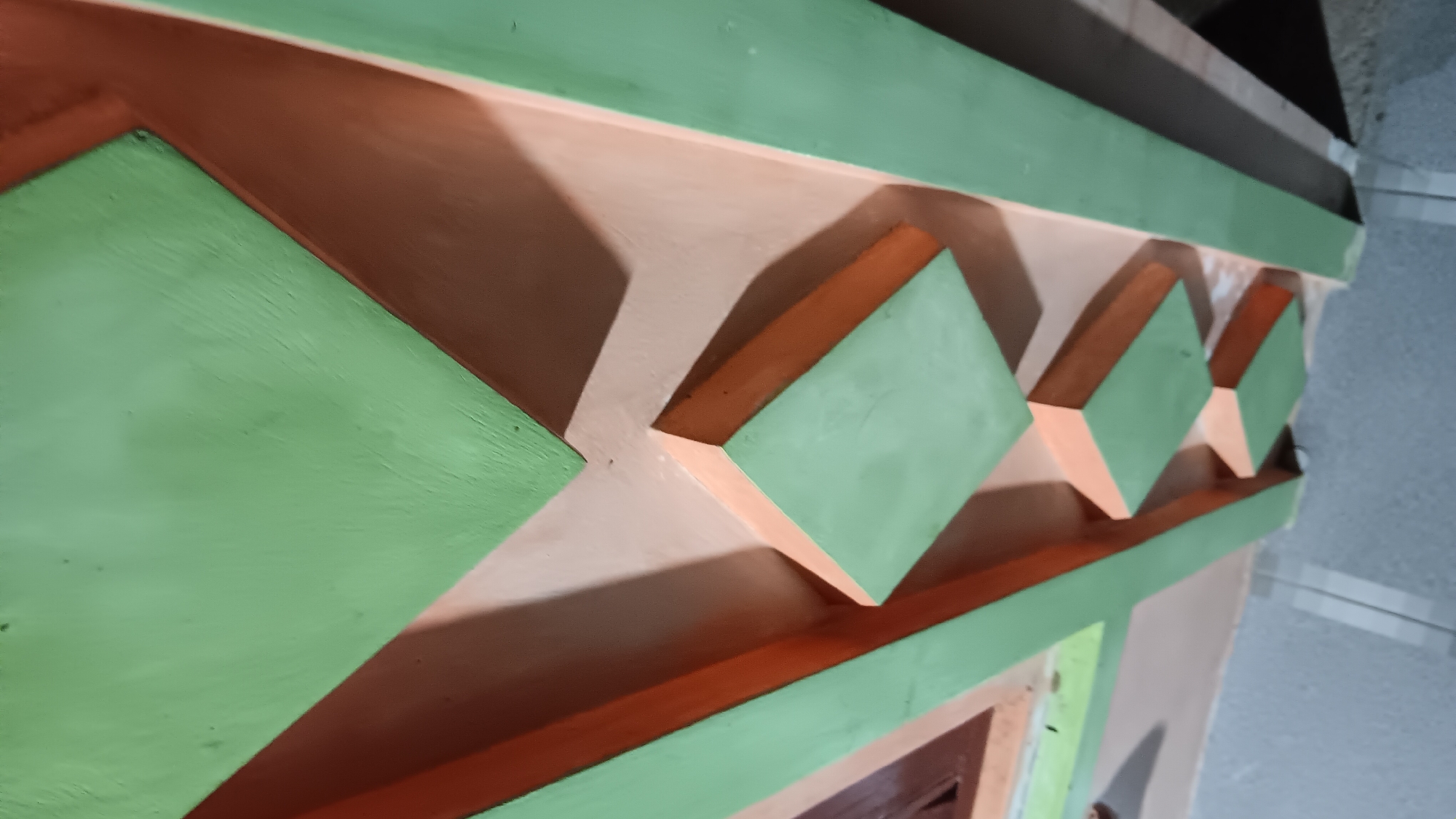 The image shows a close-up, angled view of an architectural detail, likely a decorative element on a wall or ceiling. The design features geometric shapes in intersecting planes, painted in two distinct color schemes: a light green and an orangey-brown, with a neutral beige background. These colors create a sense of depth and dimension, with shadows playing a significant role in defining the edges and forms of the shapes.
Specifically, there are several angled surfaces painted in light green, forming diamond-like or triangular shapes. These are framed and connected by elements painted in orangey-brown, which also create recessed areas and protruding edges. The texture of the painted surfaces appears somewhat rough or matte.
The context of the location in Sonobekel, Indonesia, is not visually apparent from the image itself. The image focuses solely on the abstract architectural design. No people, actions, or interactions are visible. The environment is implied to be indoors or a covered outdoor space due to the lighting and the presence of what looks like a tiled floor in the lower portion of the image.