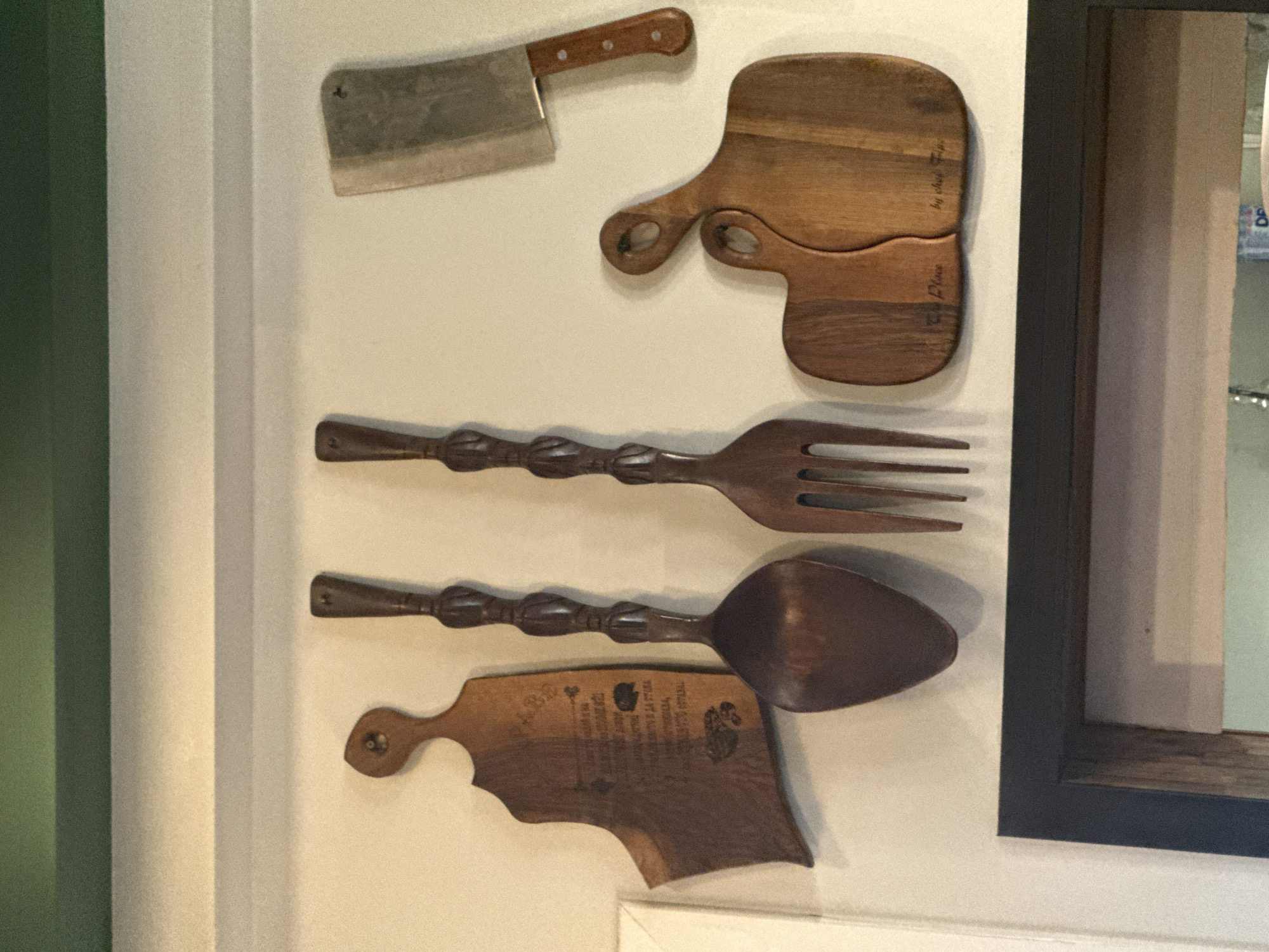 A wall display features a butcher knife, two wooden cutting boards, and a large wooden fork and spoon. The butcher knife is at the top left. Below the knife is a cutting board made of two interlocking pieces. Further down, the fork and spoon are horizontally positioned. Below the utensils is a shaped cutting board. The items are mounted on a white wall. There are architectural elements suggesting an interior location, with a green section on the left and a dark-framed element, possibly a window or display case, on the right. The location is unknown as the city of Sofia, Bulgaria, cannot be verified from the image.