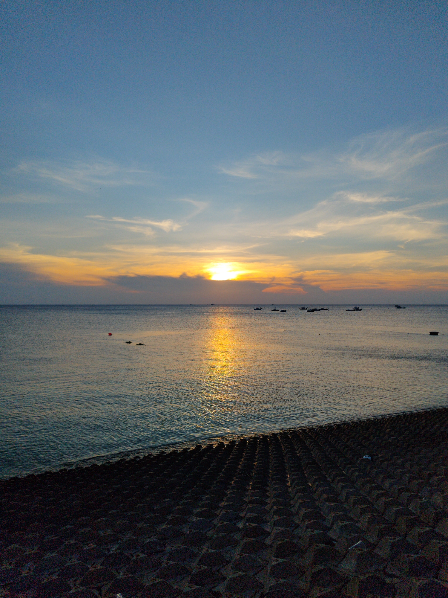 The image captures a sunset scene in Phú Quý Special Administrative Region, Vietnam. The sky transitions from blue to orange, with the sun obscured by clouds. The sun's reflection creates a bright path across the ocean's surface. Several small boats are scattered along the horizon. In the foreground, a structure consisting of rows of dark, concrete tetrahedral blocks extends into the water.
