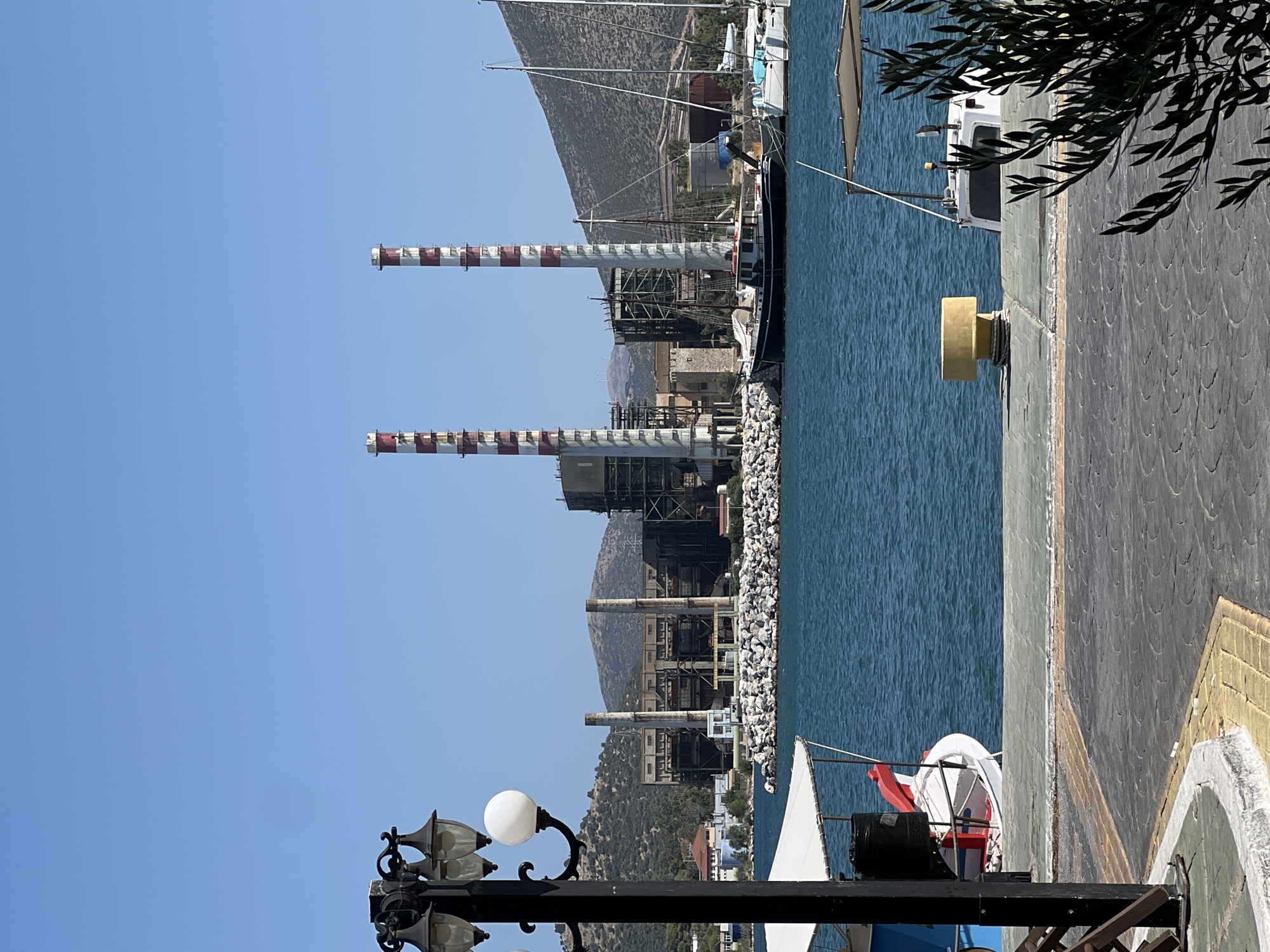 The image depicts a waterfront scene in Karavos, Greece. In the background, a large industrial facility with two tall, red and white striped smokestacks dominates the landscape. The facility appears to be a power plant or similar industrial complex, with various buildings and metal structures visible. Hills covered in sparse vegetation rise behind the industrial site.
In the foreground, a body of blue water, likely the sea, is calm with gentle ripples. Several boats are docked in the harbor. Two sailboats are prominent, one with a white hull and mast, and another dark-hulled vessel with sails furled. A smaller white boat with a red and white trim is also visible.
The waterfront itself is paved with grey interlocking stones, with a yellow-lined border. A black lamppost with multiple lights stands on the left side of the image, partially obscuring a white canopy over a boat. A yellow bollard is positioned on the pier near the water's edge. Olive tree branches are visible in the lower right corner, framing the scene. The sky is clear and blue, indicating fair weather. There are no people visible in the image.