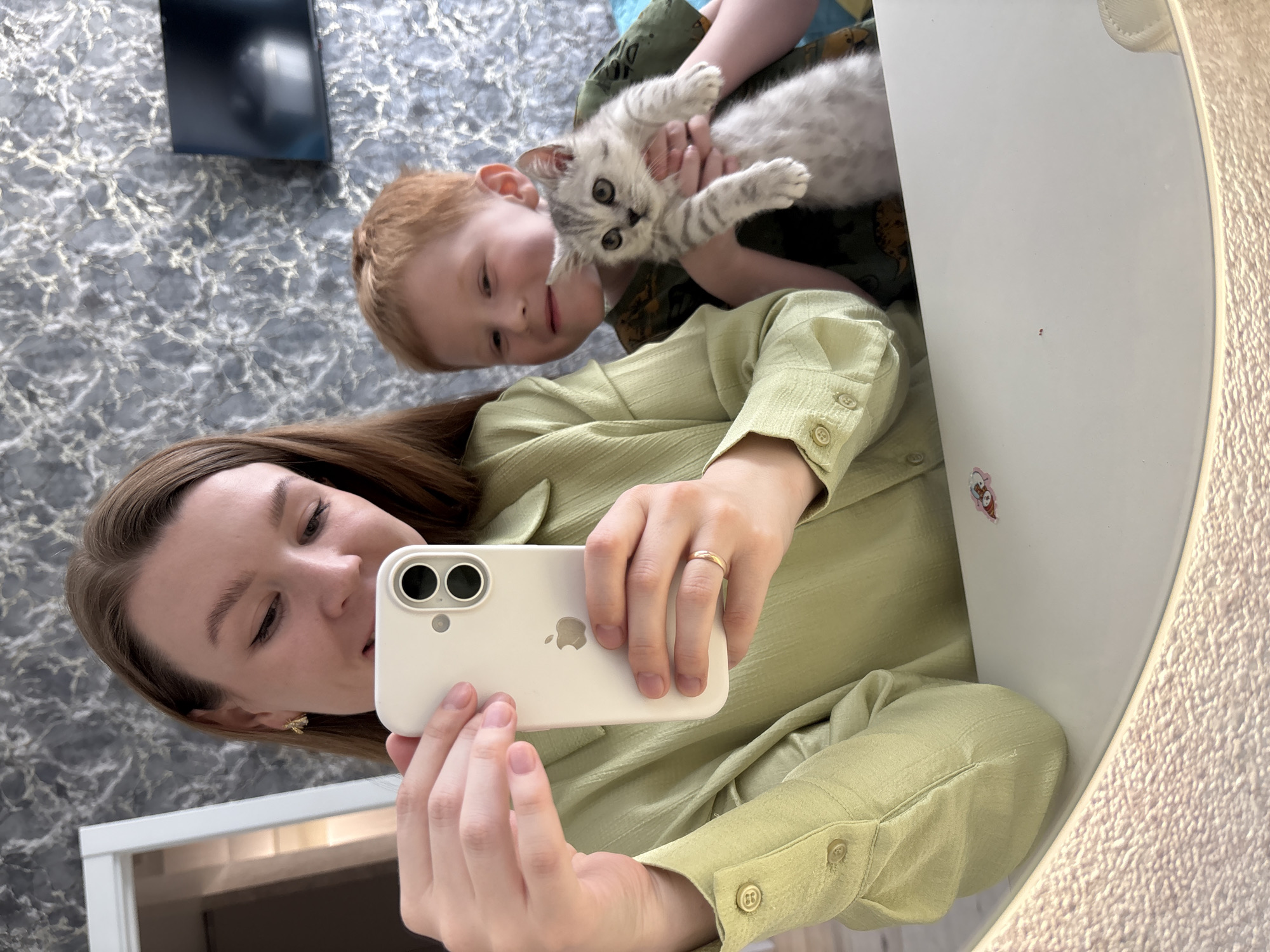 A woman is taking a mirror selfie with a child and a kitten. The woman is holding a white smartphone and wearing a light green shirt. The child, who appears to be a young boy with red hair, is holding a grey tabby kitten. The background shows a bathroom with a grey and white wall and a white doorframe. There is a television mounted on the wall. It is not possible to determine the location within Izhevsk, Russia from the information given in this picture.