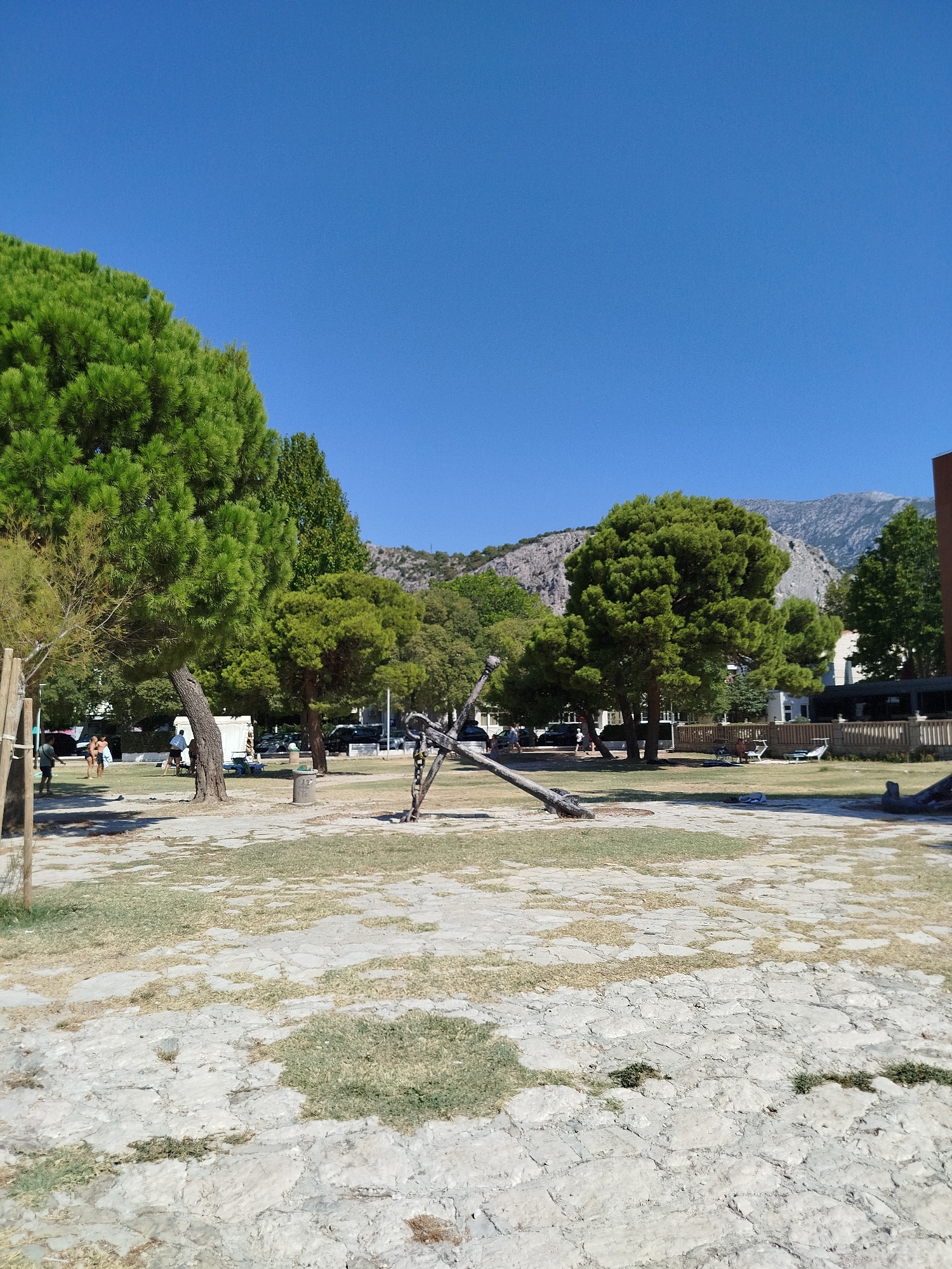 This is a vertical, eye-level, outdoor shot of a paved area with scattered grass, large pine trees, and a clear blue sky. A large, dark-colored anchor is the main subject, situated in the center of the frame, propped up by its flukes. In the background, there are more trees, buildings, a wooden fence, parked cars, and a mountainous landscape under the bright sun. A few people are visible, scattered around the park, with one person walking on the left side of the frame and others sitting on lounge chairs near the fence. The ground is covered in large, unevenly shaped light-colored stones, with patches of dry and green grass. The overall atmosphere is one of a sunny day in a park.