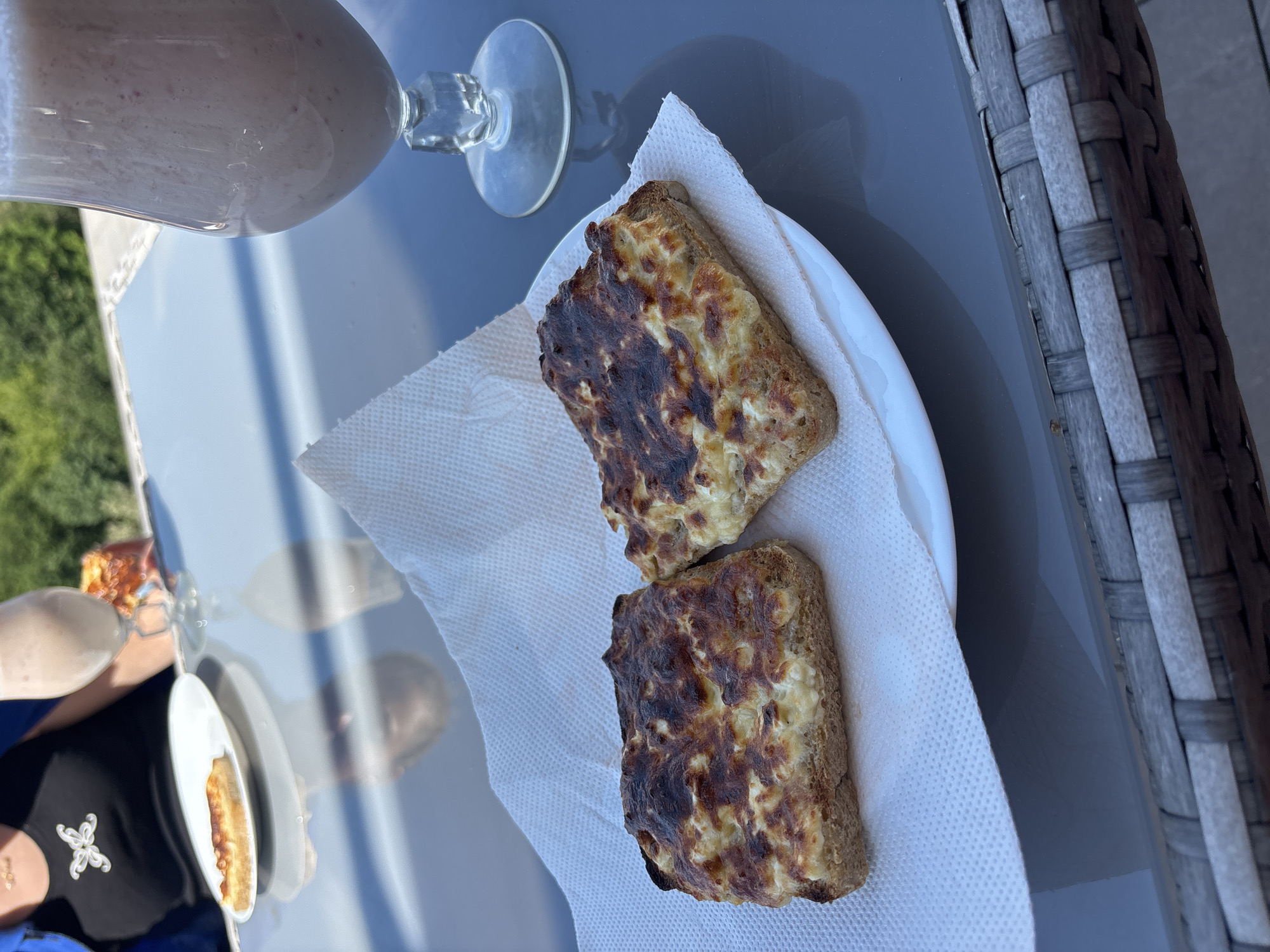 The scene is an outdoor setting, possibly a patio or balcony, in daytime. A grey surface, likely a table, holds a partially filled glass with a light pink smoothie and a white plate containing two pieces of toast with a melted topping that appears browned and slightly burnt. The toast rests on a white paper napkin. To the left, a reflection on the table shows another person sitting at the table with a black shirt with a small white flower, and a plate with a sandwich with melted topping on it. A woven grey structure, perhaps part of outdoor furniture, is seen along the right edge of the image. The green vegetation and blue sky are visible through reflection in the table. The location is Tserovo, Bulgaria.