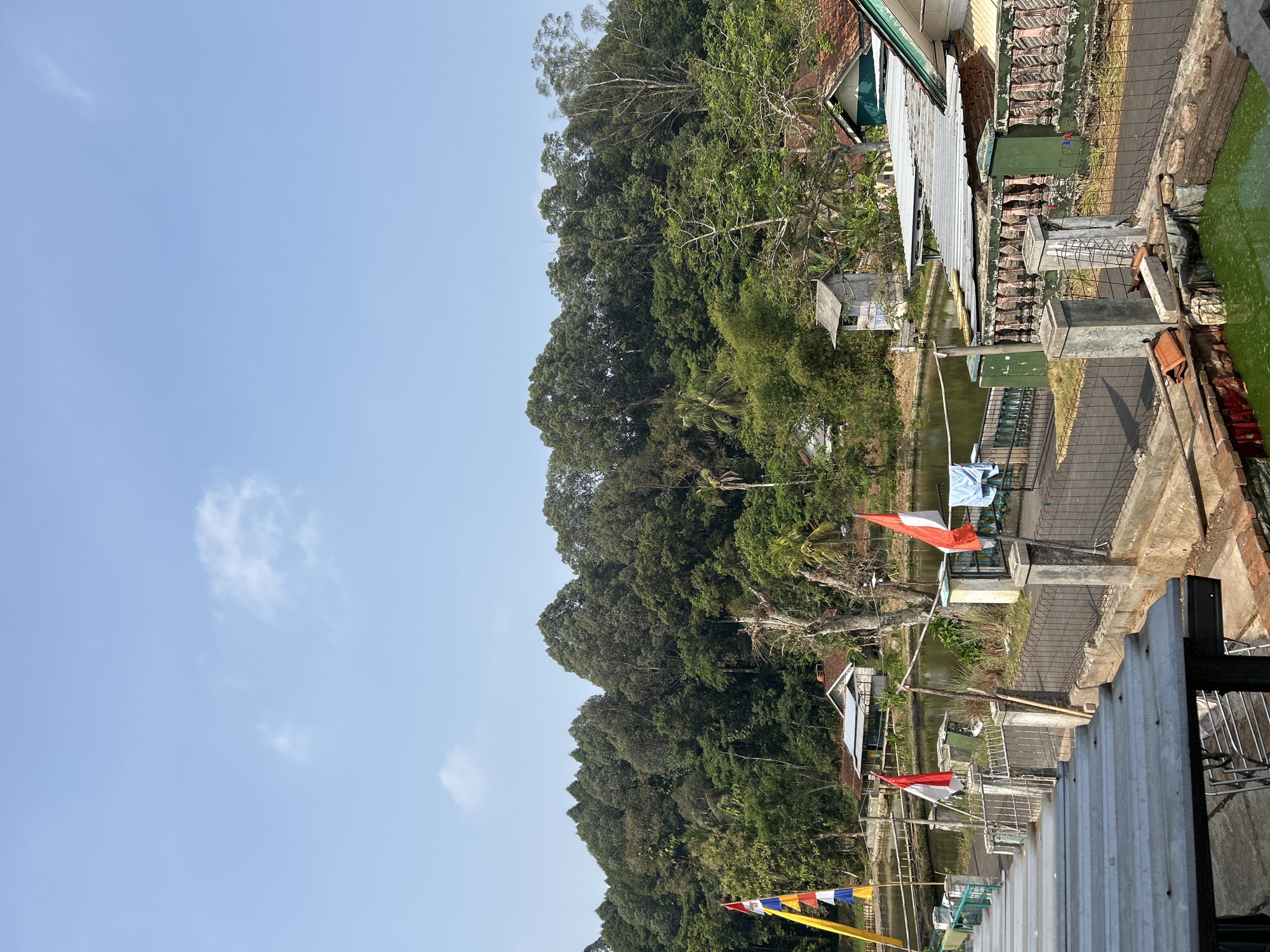 The image shows a view of a small village in Cisaga, Indonesia. In the foreground, a corrugated metal roof is visible, suggesting a covered walkway or building. A paved road runs through the scene, bordered by a low fence with decorative stone pillars.

Several flags are displayed, including Indonesian flags with red and white bands, and a yellow and blue striped flag. There are small buildings with tiled roofs, and a body of water, possibly a pond or lake, is present. The background is dominated by a lush, green hillside covered in dense trees. Laundry hangs on a clothesline between two flags. The sky is clear blue with a few wispy clouds. The scene appears to be a rural or semi-rural setting in the daytime.