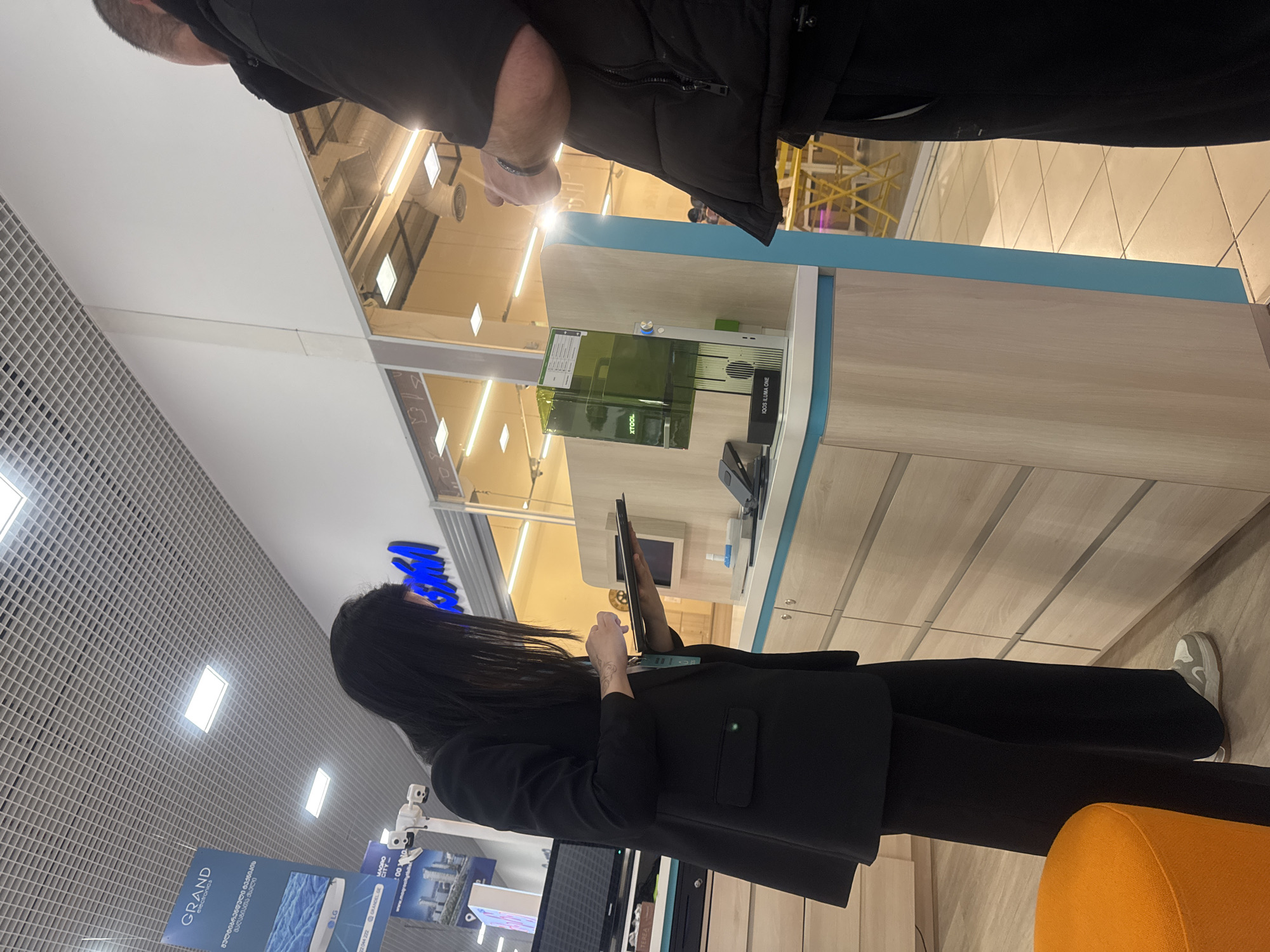 This image depicts an indoor scene within a retail environment, likely an electronics store or a dedicated brand kiosk, situated in Tbilisi, Georgia.
In the foreground, a woman with long dark hair, wearing a black blazer and trousers, is standing and holding a tablet. She appears to be interacting with the device. A tattoo is visible on her left forearm. Partially visible in the upper part of the frame is a man, seen from the chest up, wearing a dark jacket. He is positioned opposite the woman, possibly observing or waiting.
The central feature of the setting is a modern counter with light wood and blue trim. On this counter, a transparent green-tinted device is displayed, labeled "XTOOL", alongside another product distinctly marked "IQOS ILUMA ONE" and a small "0010". A bottle of hand sanitizer is also on the counter.
The background reveals a retail space with a grid-patterned ceiling featuring square lighting fixtures. Prominent signs are visible, including "GRAND electronics" which also displays the "LG" logo. Beneath "GRAND electronics," Georgian text reads "მაღაზიათა ქსელი" (Store Network) and "მულტიბრენდული ტექნიკის" (Multi-brand appliances), along with contact information "22044 222 GRANDEL GRO". An orange upholstered chair is visible in the lower right corner, suggesting a waiting or consultation area. The overall lighting is bright and artificial, typical of an indoor commercial space.